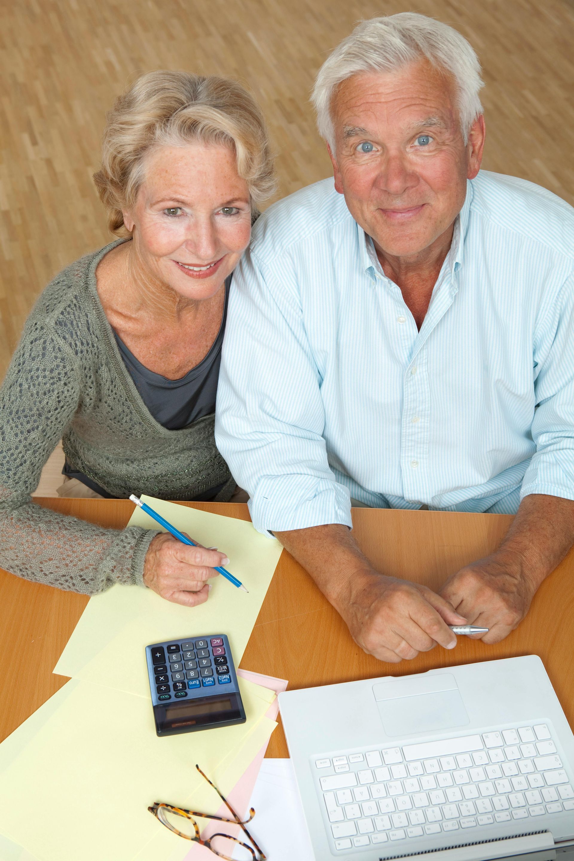 Two people at a desk reviewing paperwork with a calculator and keyboard, looking up at the camera.