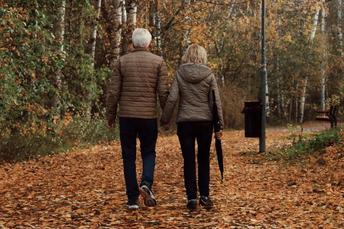 Two people walking away on a leaf-covered forest path in autumn, one holding a cane