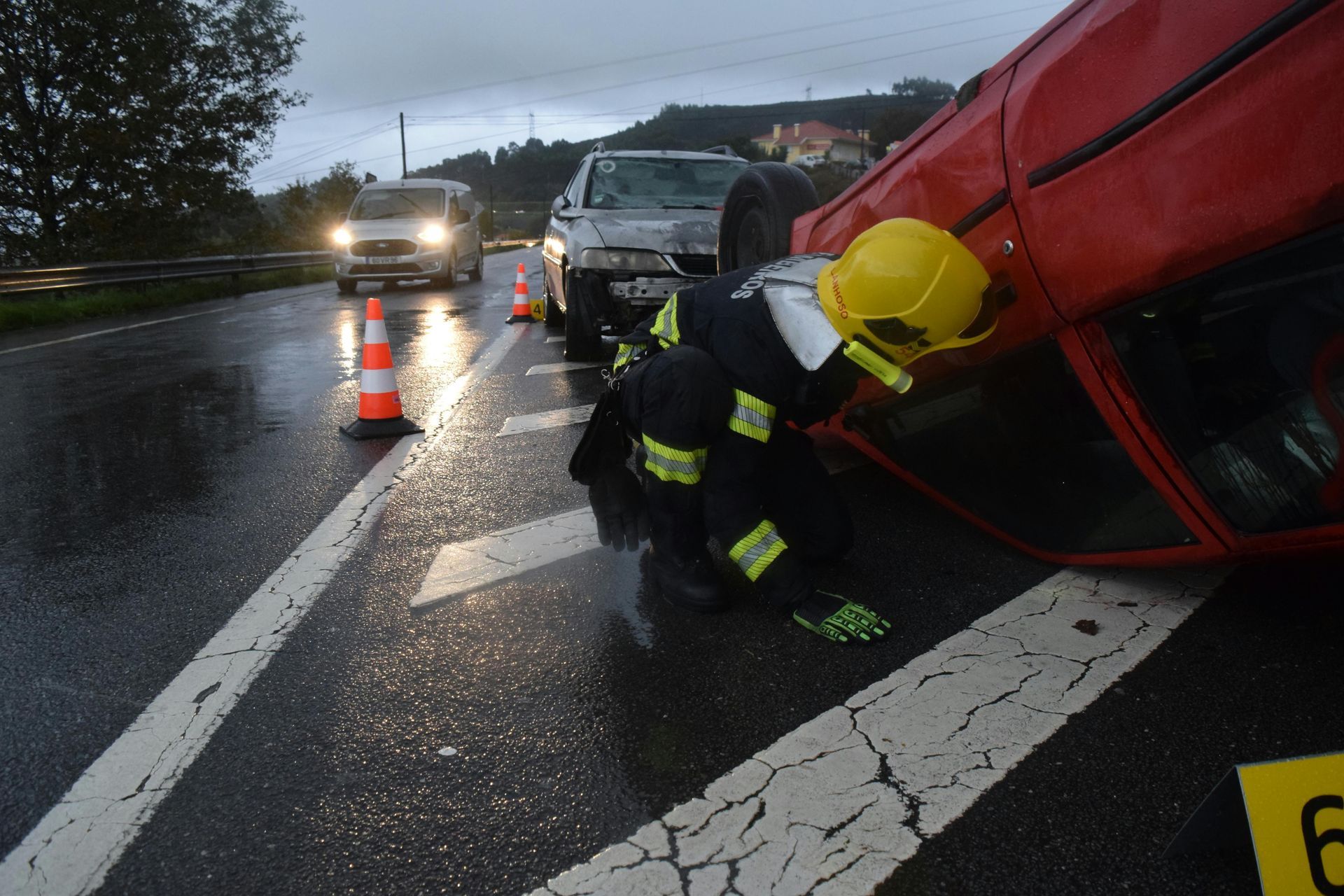 Firefighter in yellow helmet rescues at a rainy roadside crash with an overturned red vehicle.
