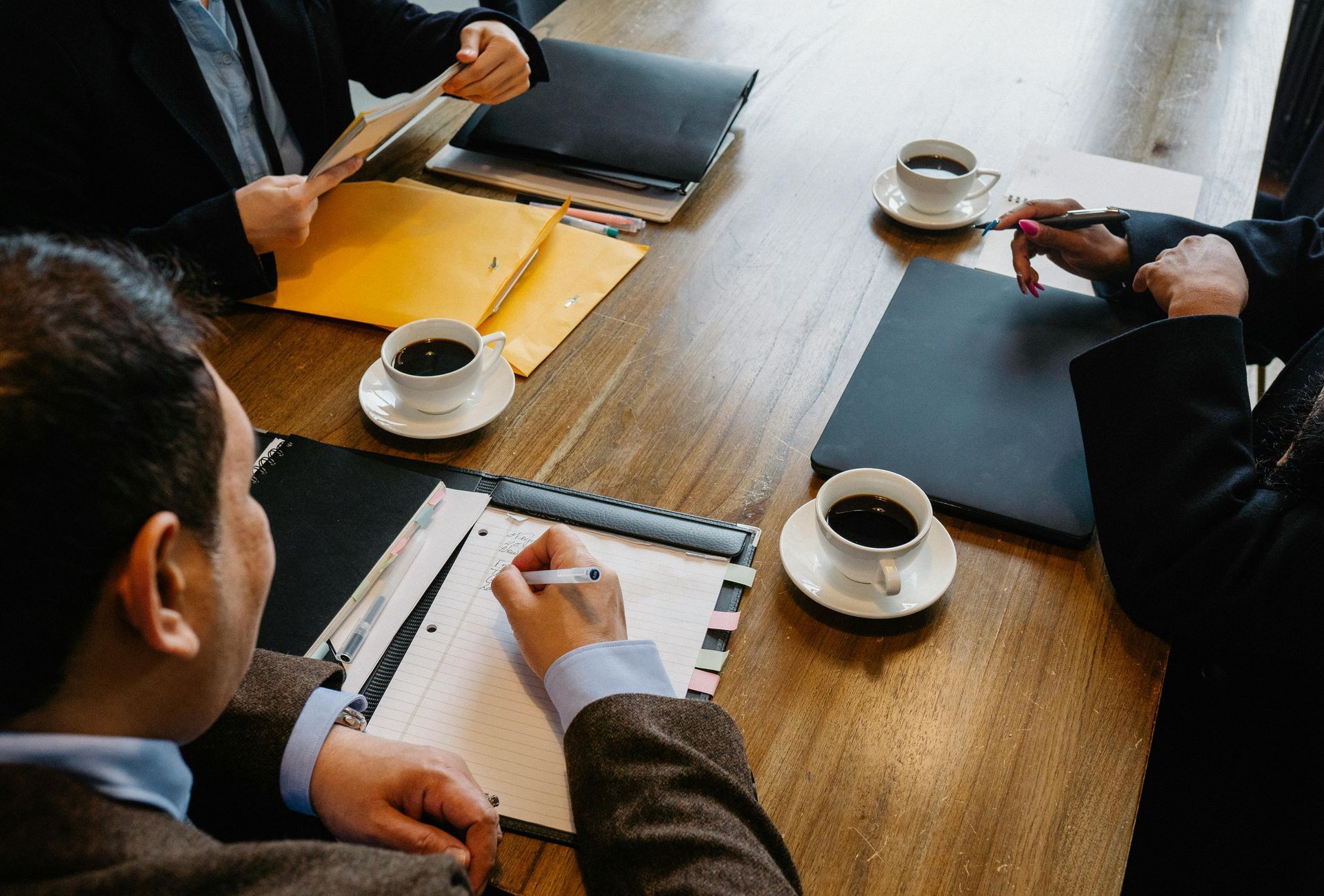 People in a meeting around a wooden table with coffee cups and notebooks, discussing documents.