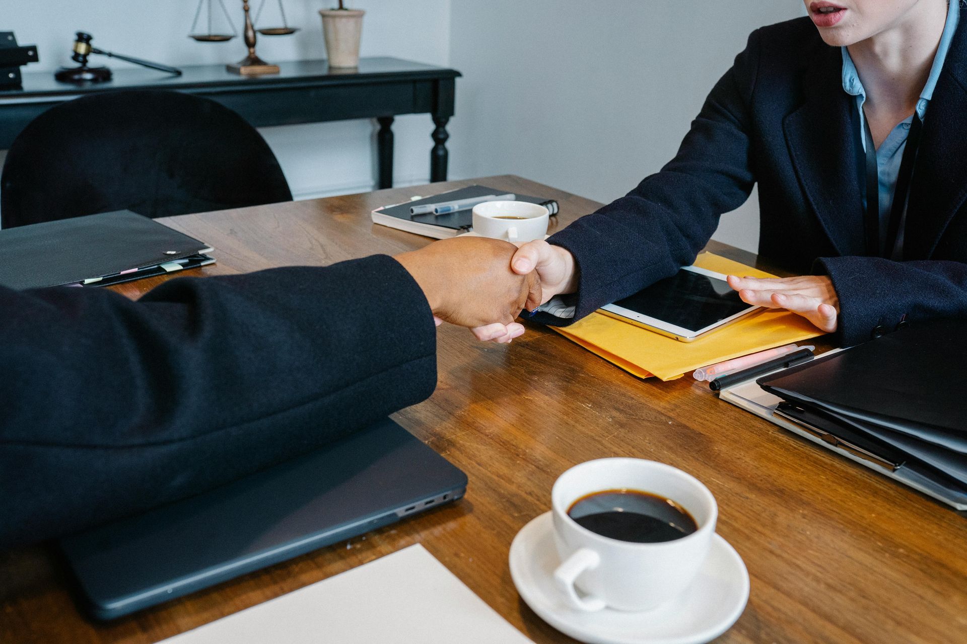 Two people shaking hands across a wooden meeting table with coffee and folders nearby