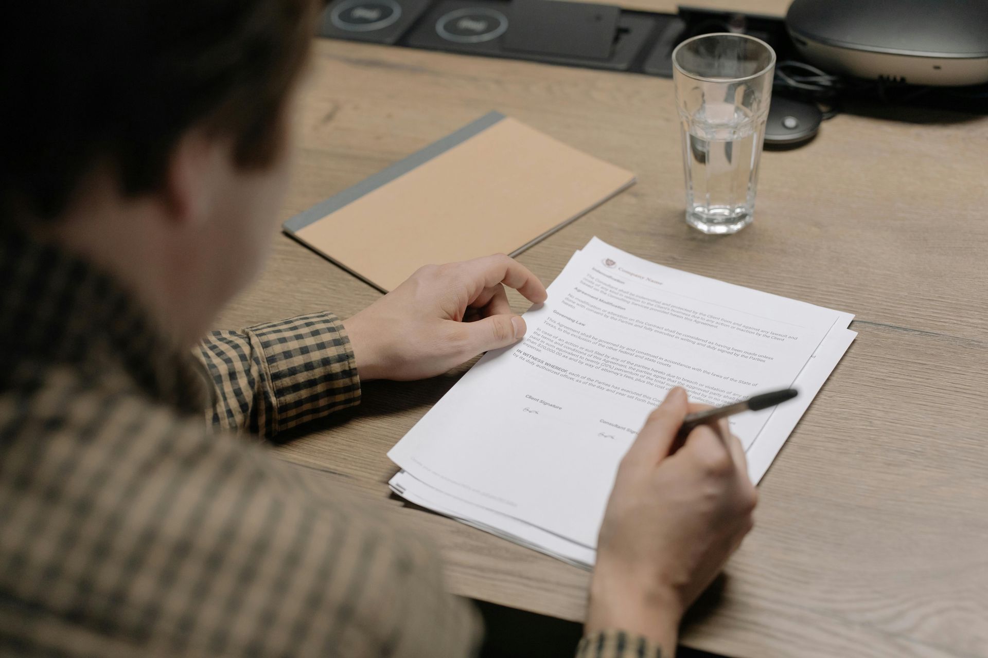 Person filling out paperwork at a desk with a pen, with a glass of water and notebook nearby.