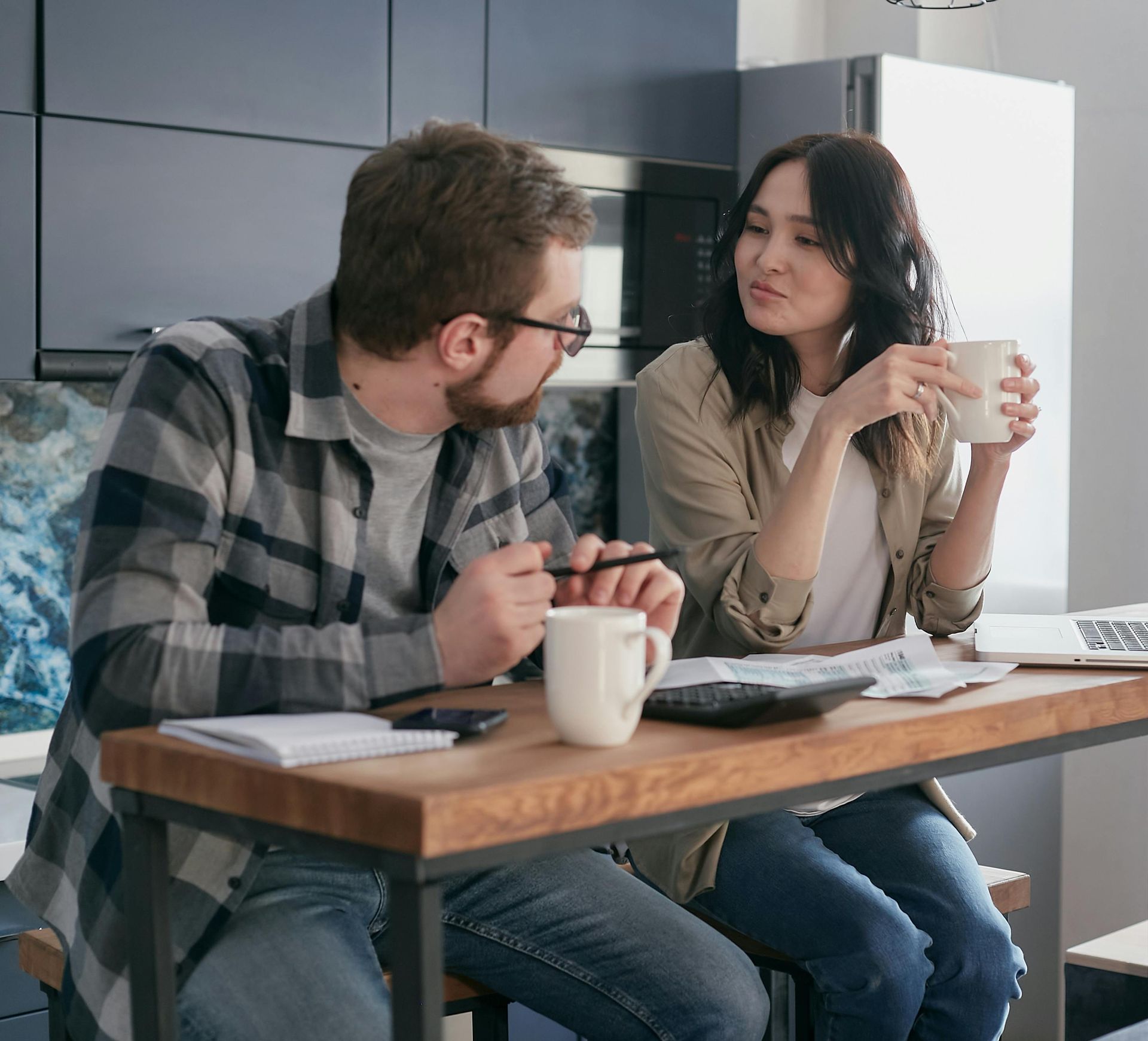 Two coworkers talking at a table with coffee mugs and a notebook in a modern office kitchen.
