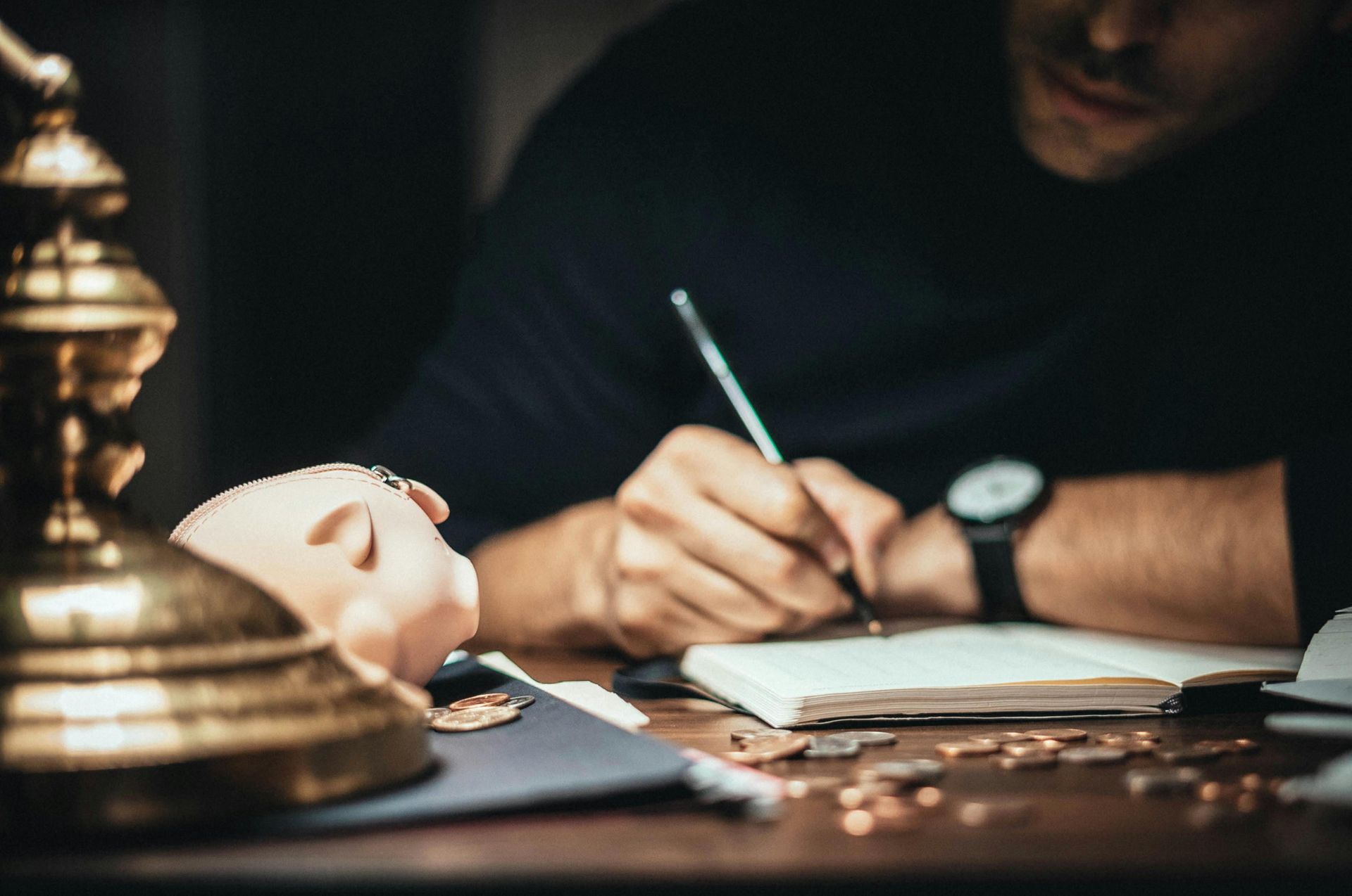 Person writing at a desk beside a brass lamp and a white mask in a dimly lit room