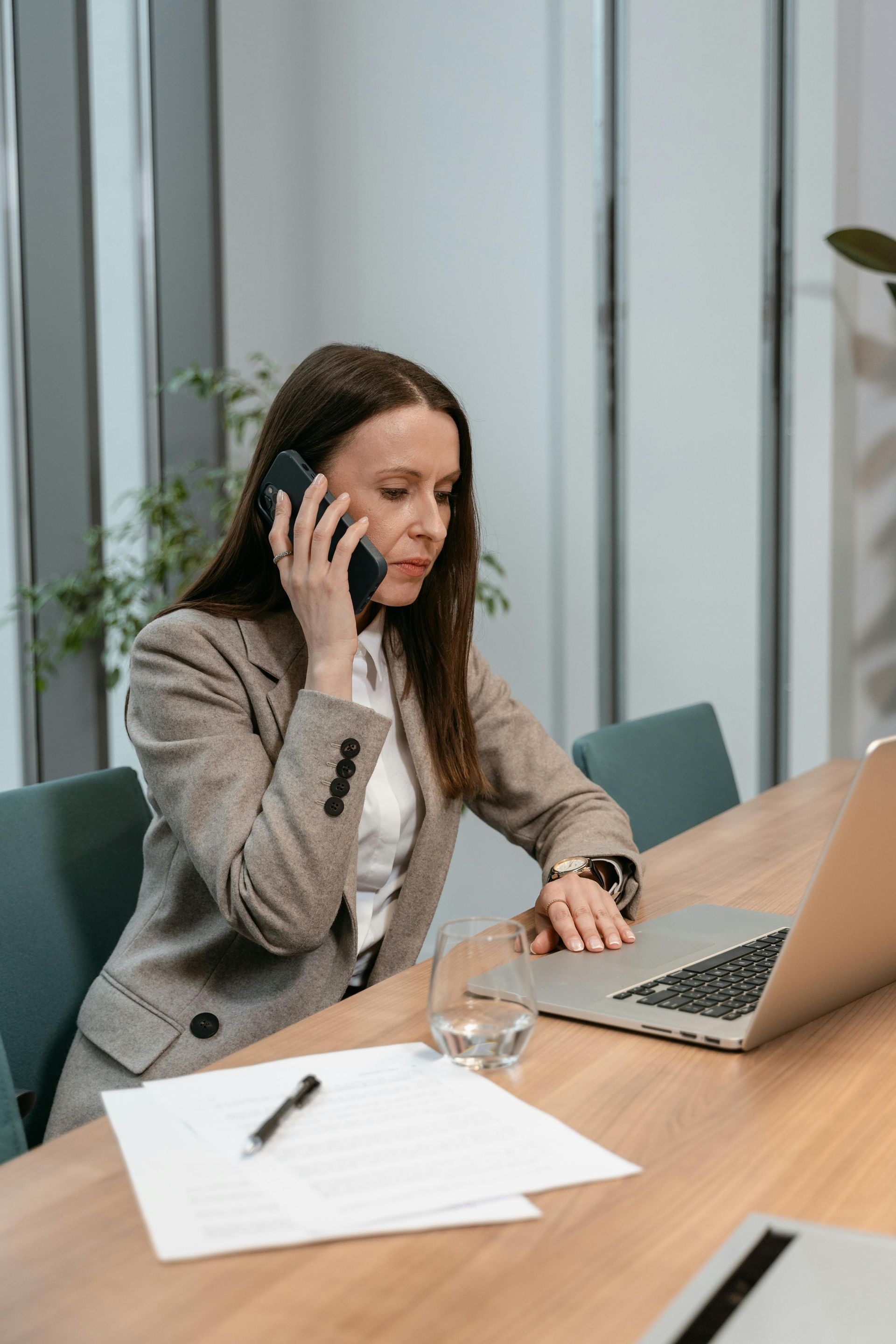 Woman on a phone call at a desk, looking at a laptop with papers and a glass of water nearby