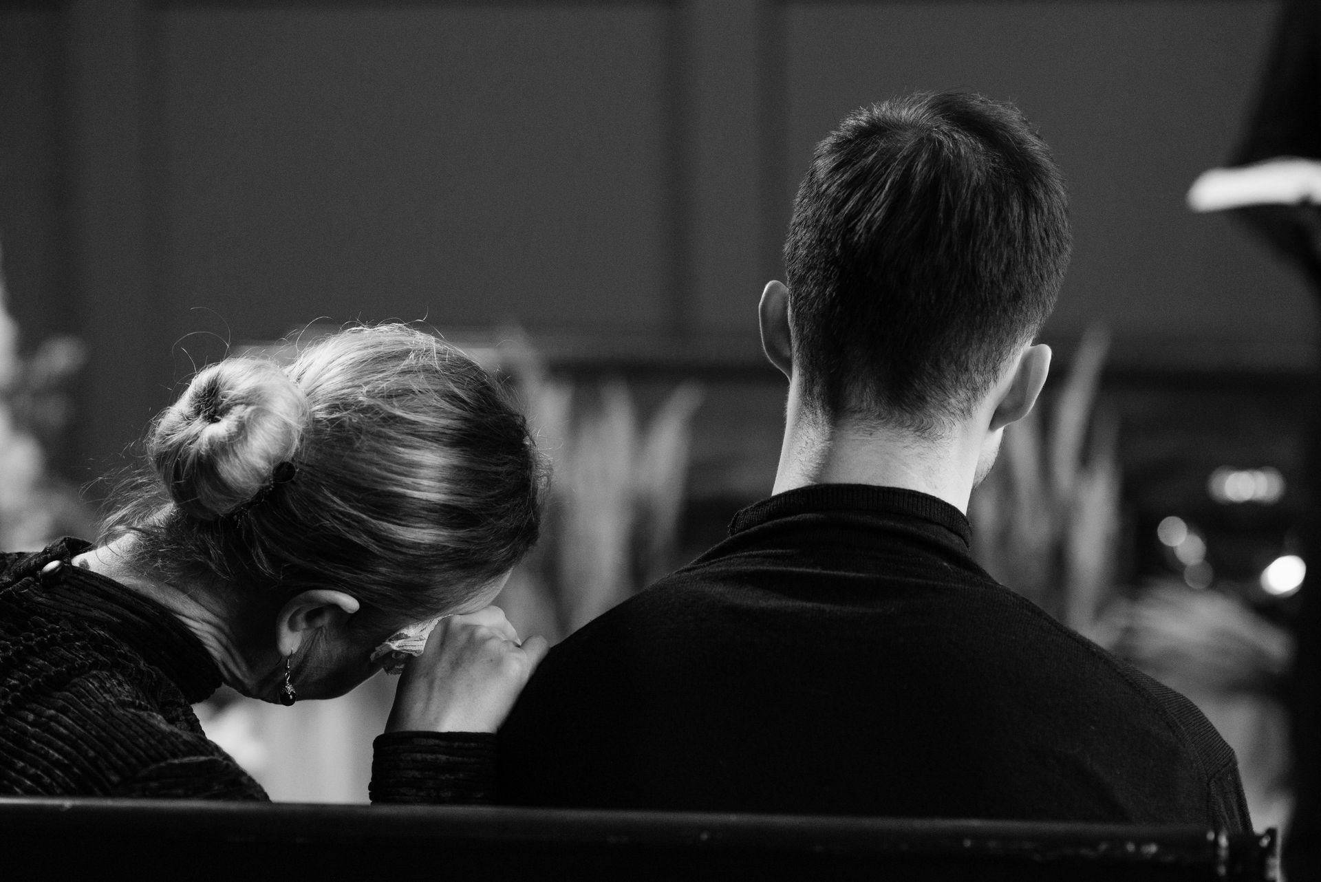 Two people seated closely; one rests their head on the other's shoulder in a dim black-and-white scene