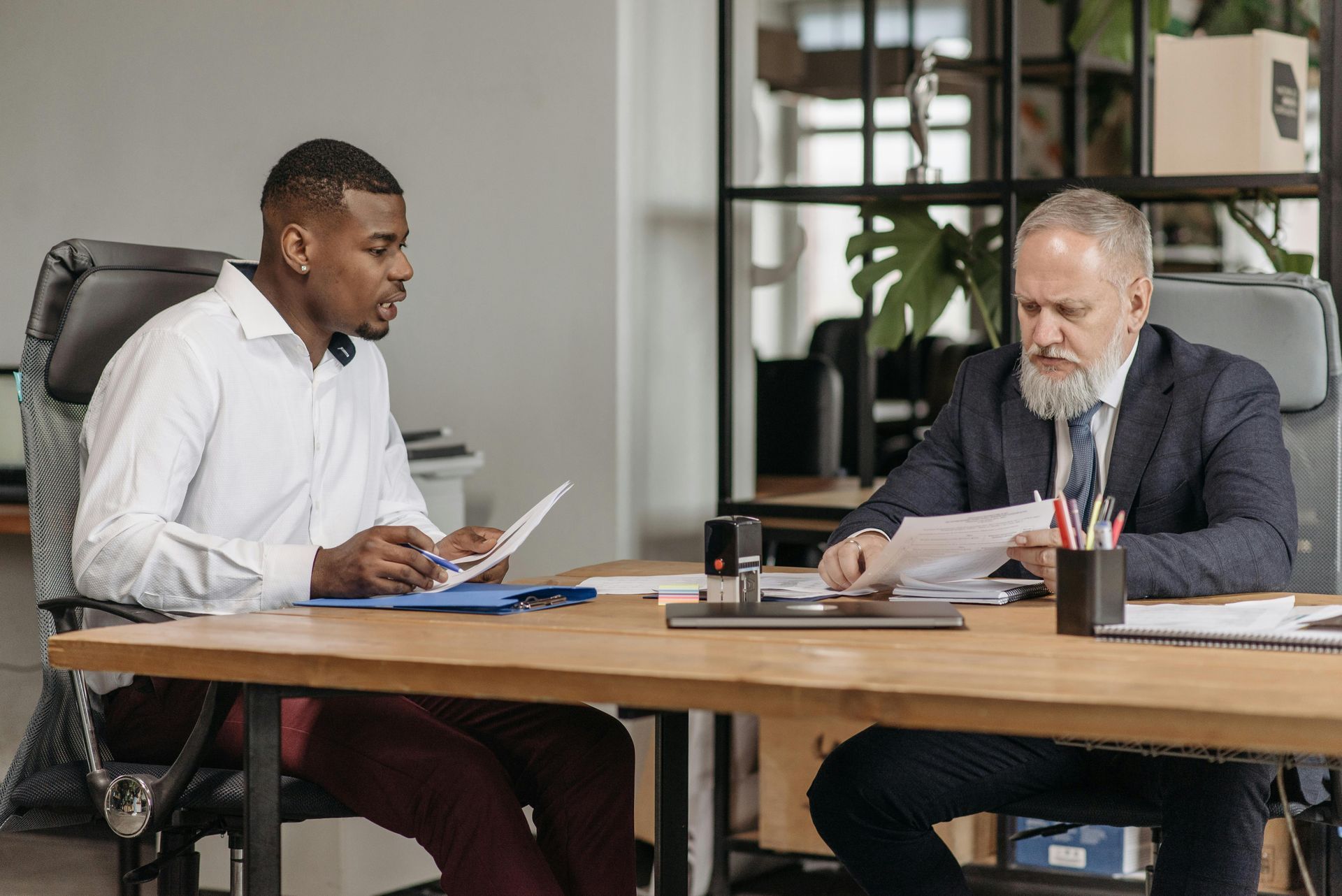 Two men discuss paperwork at a desk in an office setting.