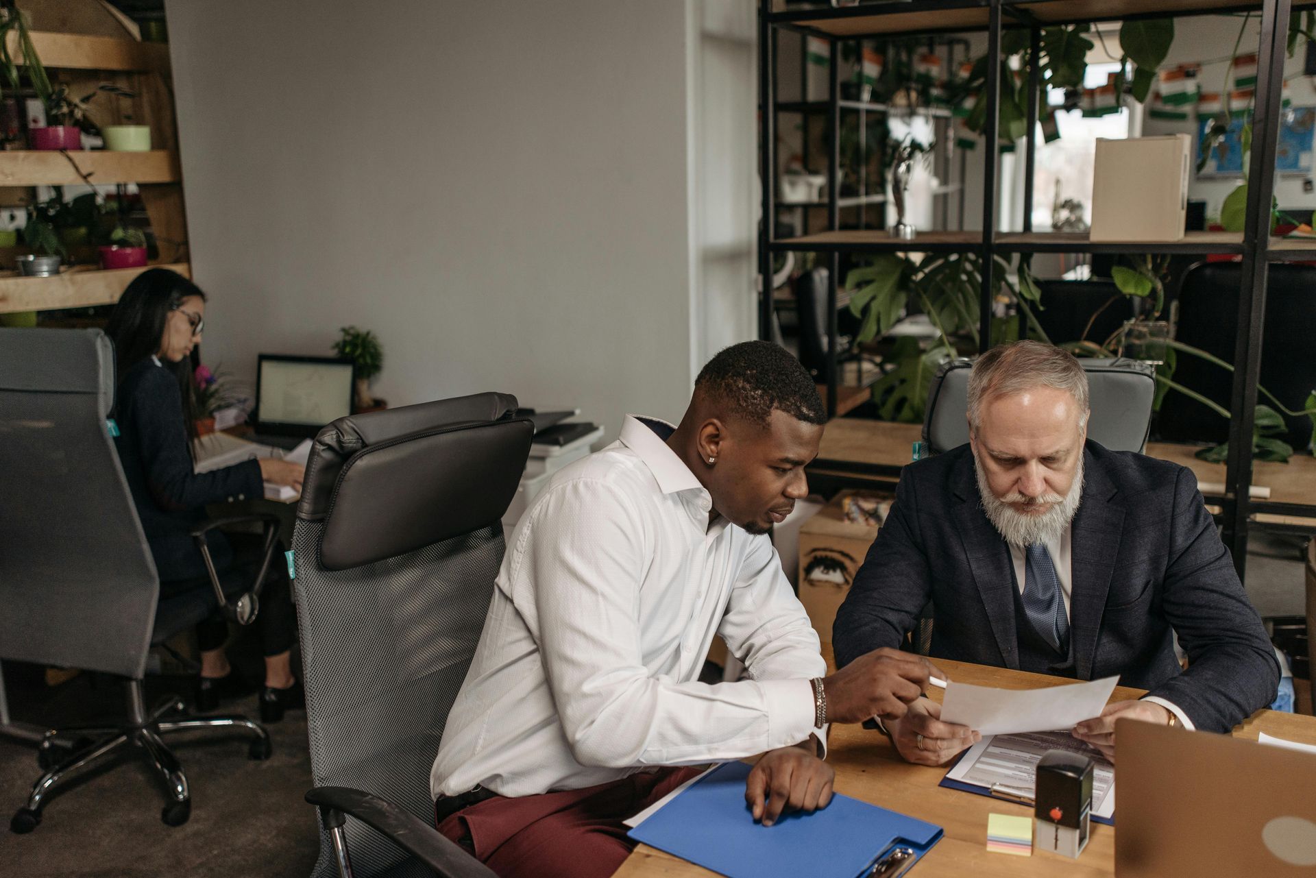 Two coworkers review documents at a desk in a modern office, with a laptop and plants nearby.