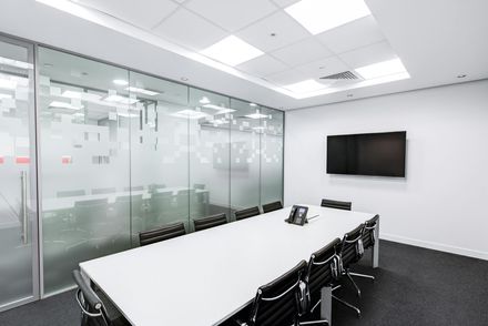 A modern, minimalist meeting room with a white conference table, black leather chairs, glass walls, and a wall-mounted TV.