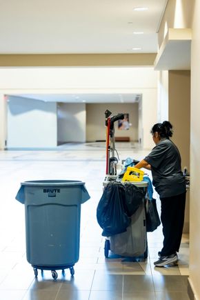 A person in a gray uniform works at a cleaning cart in a bright, spacious hallway next to a large blue trash bin.