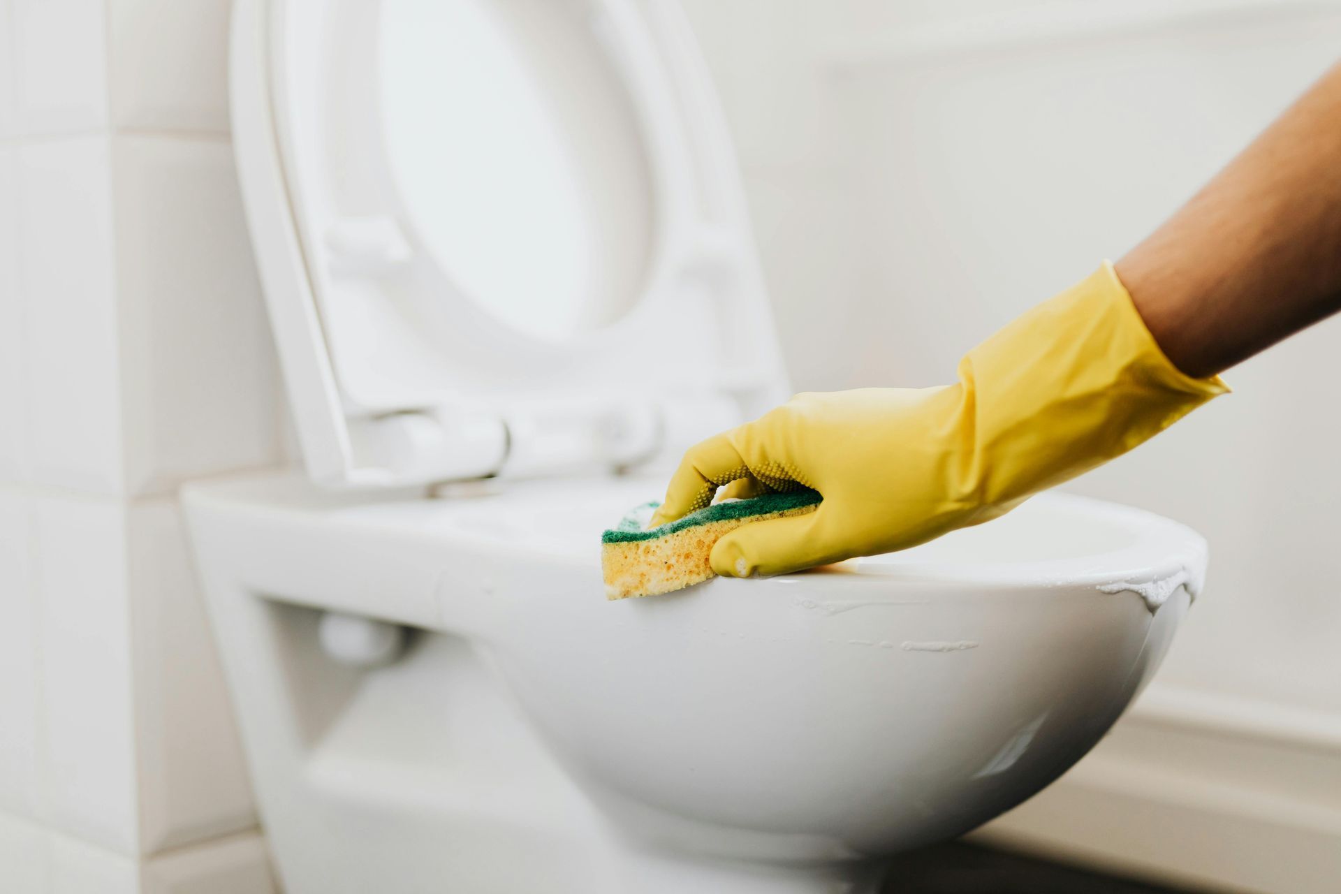 A hand in a yellow rubber glove uses a sponge to scrub the rim of a clean white toilet.