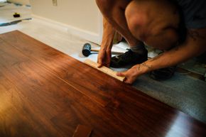 A person installs dark wood laminate flooring, using a tapping block and mallet to click a plank into place.