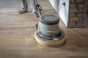 A worker uses a floor sanding machine to smooth out a wooden floor in a room under construction.