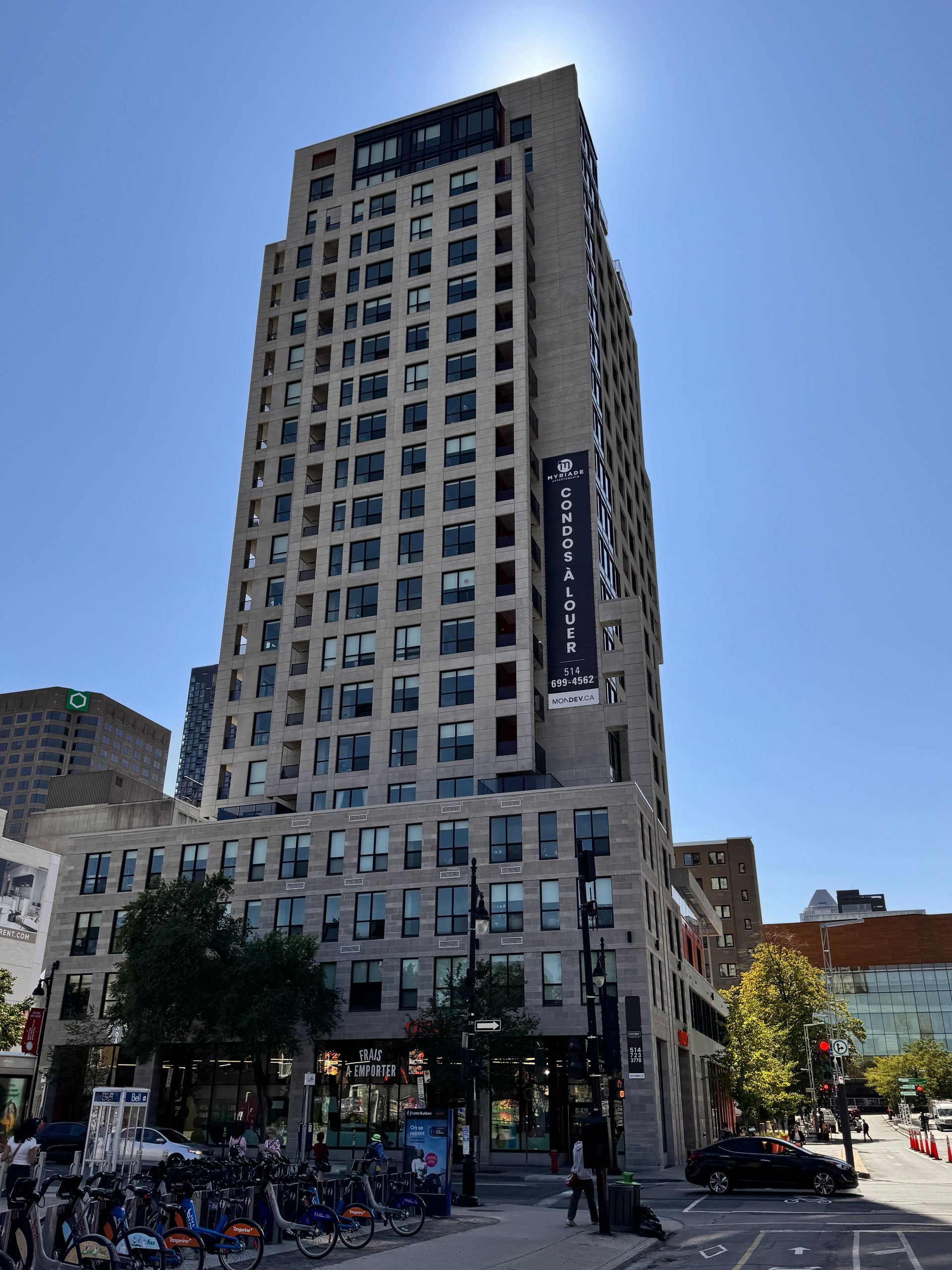 Grand immeuble d'appartements gris avec une banderole, des commerces au rez-de-chaussée, un ciel bleu et des vélos garés.