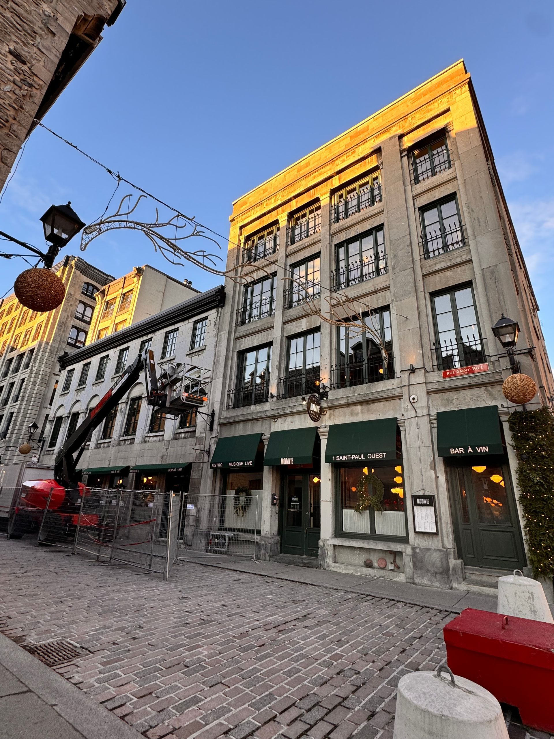 Des bâtiments en pierre bordent une rue pavée ; un restaurant aux auvents verts. 