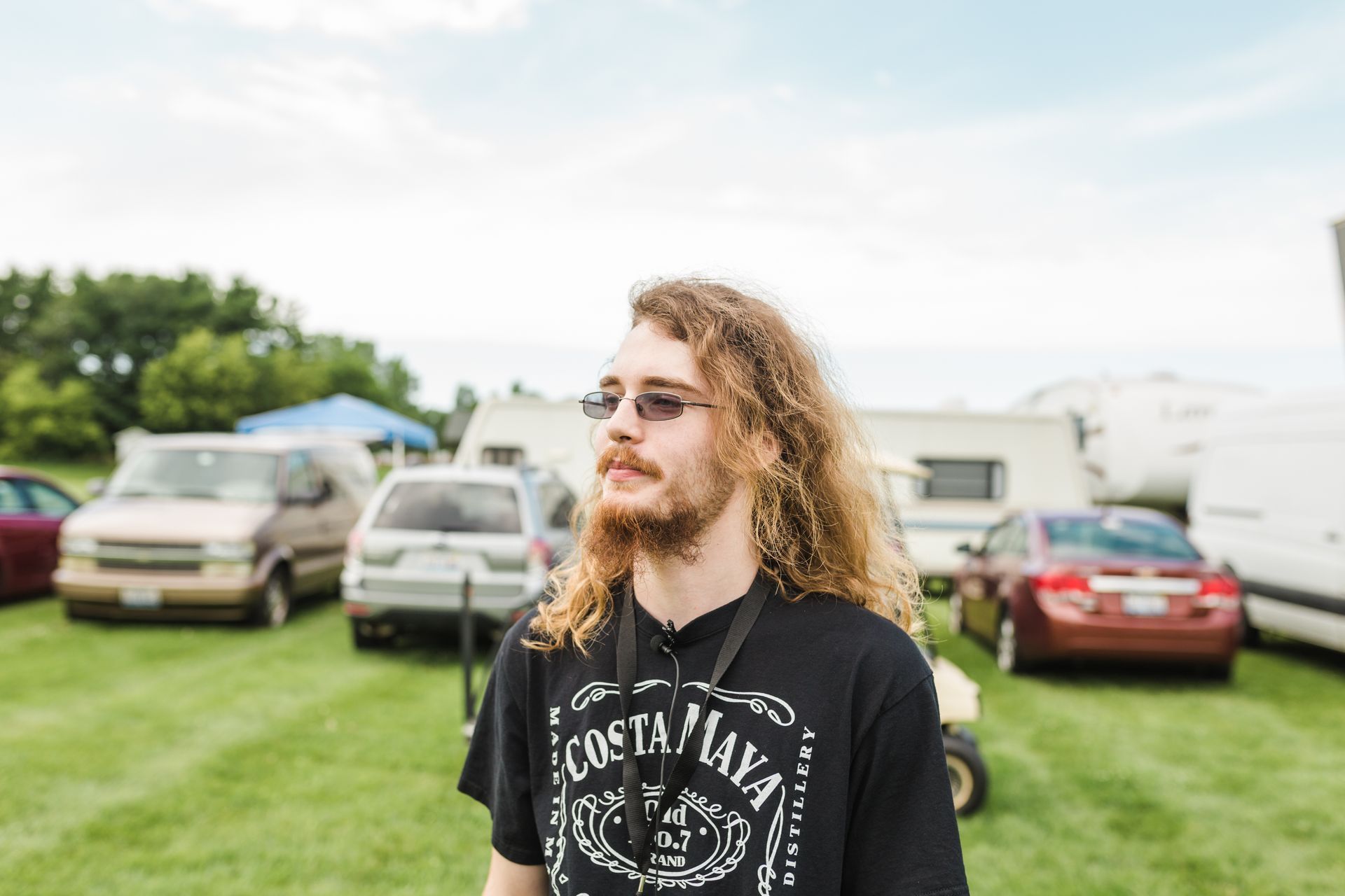 A man with long hair and a beard is standing in a field with cars parked in the background.