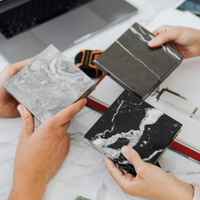 Hands comparing three dark tile samples over a white design desk with plans