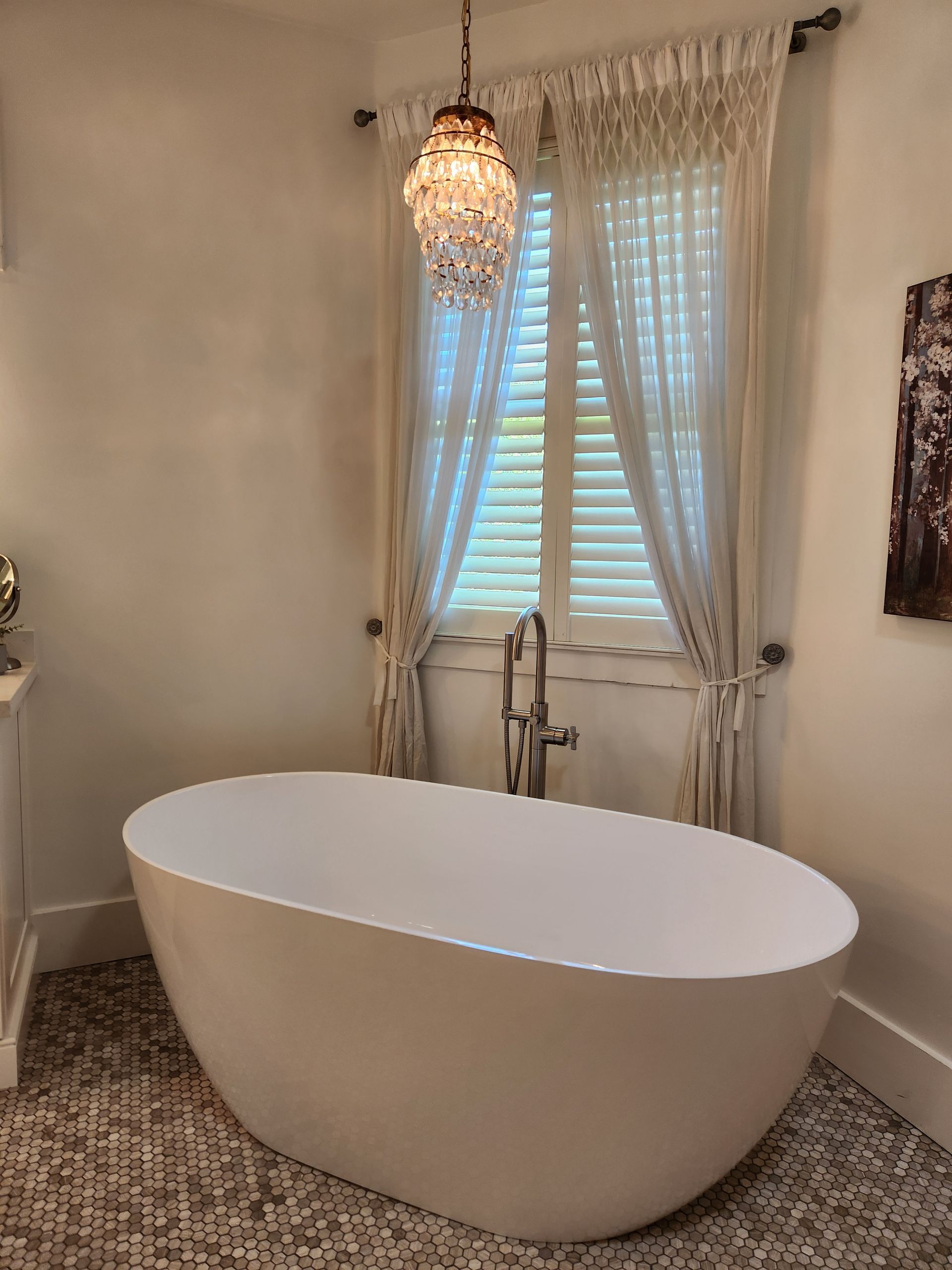 Modern freestanding oval bathtub in a bright bathroom with window, sheer curtains, and patterned tile floor