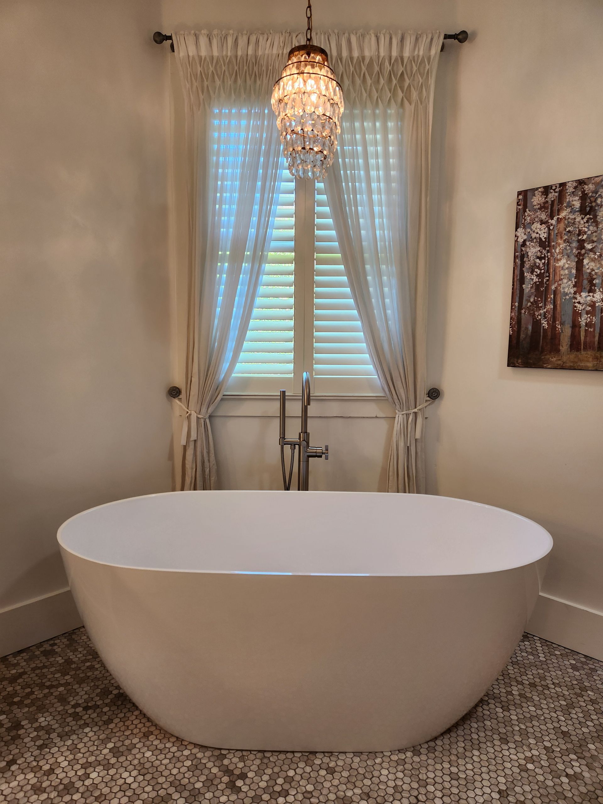 Freestanding white bathtub beneath a window with sheer curtains and a hanging chandelier in a beige bathroom