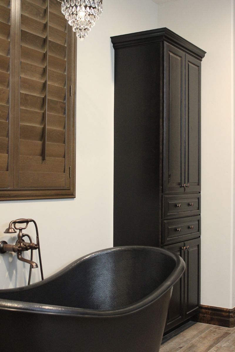Black clawfoot bathtub beside a tall dark cabinet in a bathroom with wooden window shutters and chandelier