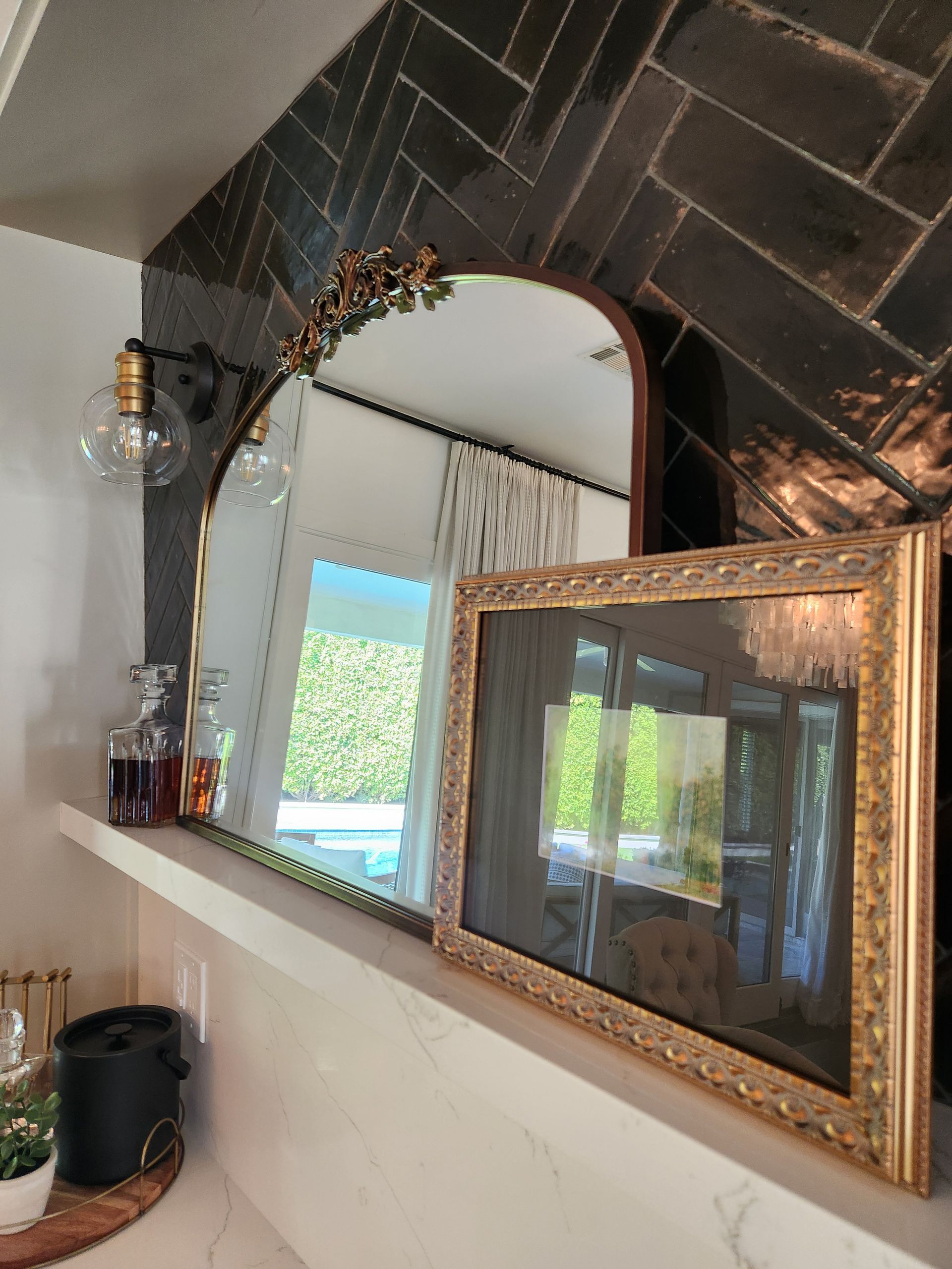 Framed mirror on a tiled wall above a shelf with a plant, bottles, and a kettle near a bright window.