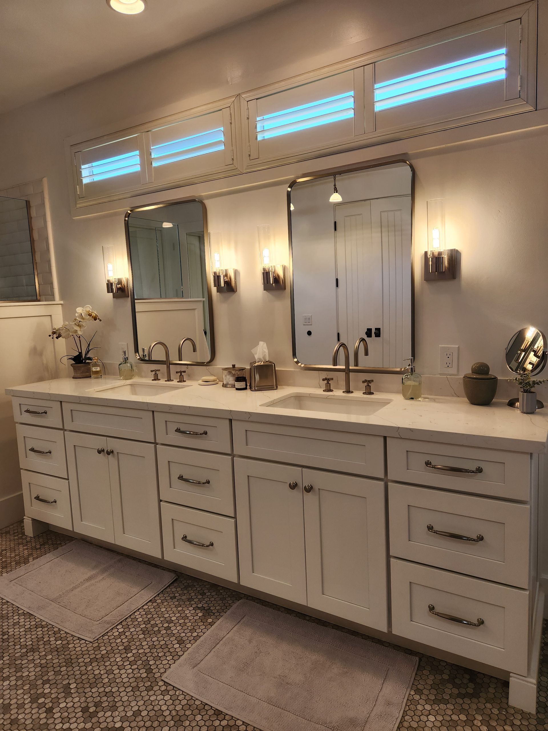 Spacious white bathroom vanity with dual mirrors, wall sconces, and marble countertop under high windows