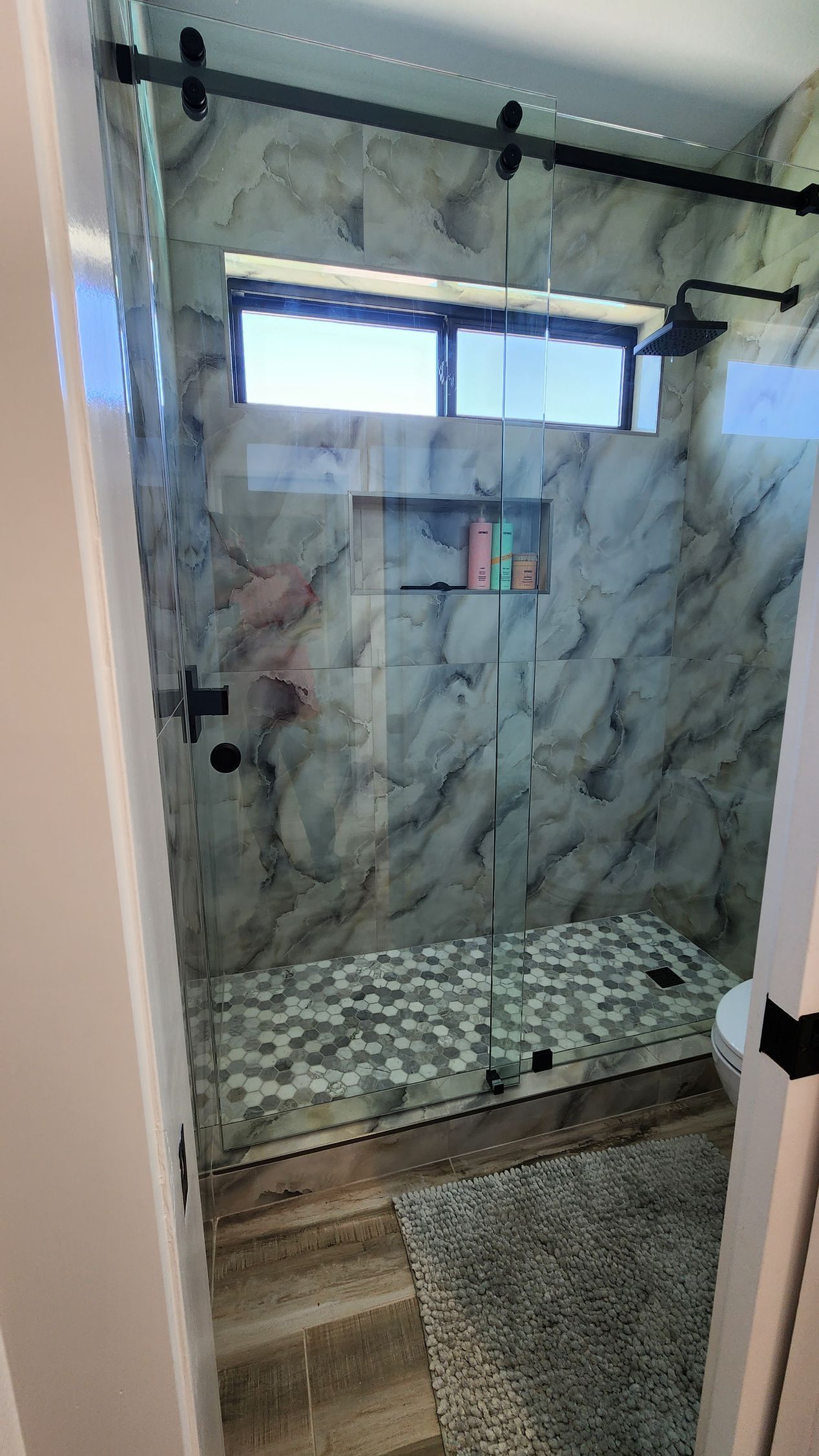Glass-enclosed shower with marble-pattern tile, frosted window, and patterned floor mat beside the door
