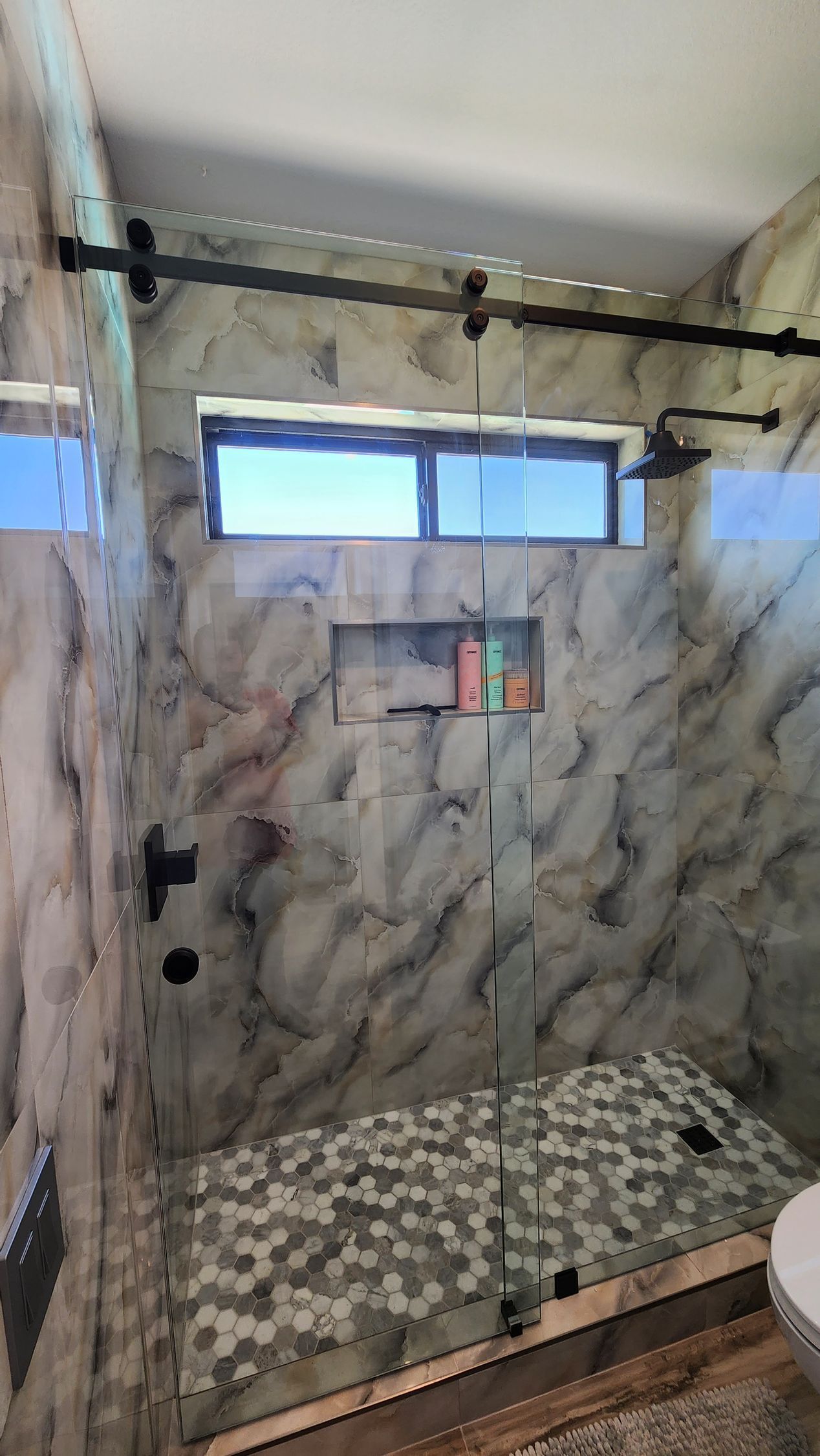 Small tiled shower stall with marble-pattern walls, a frosted window, and a black curtain rod.