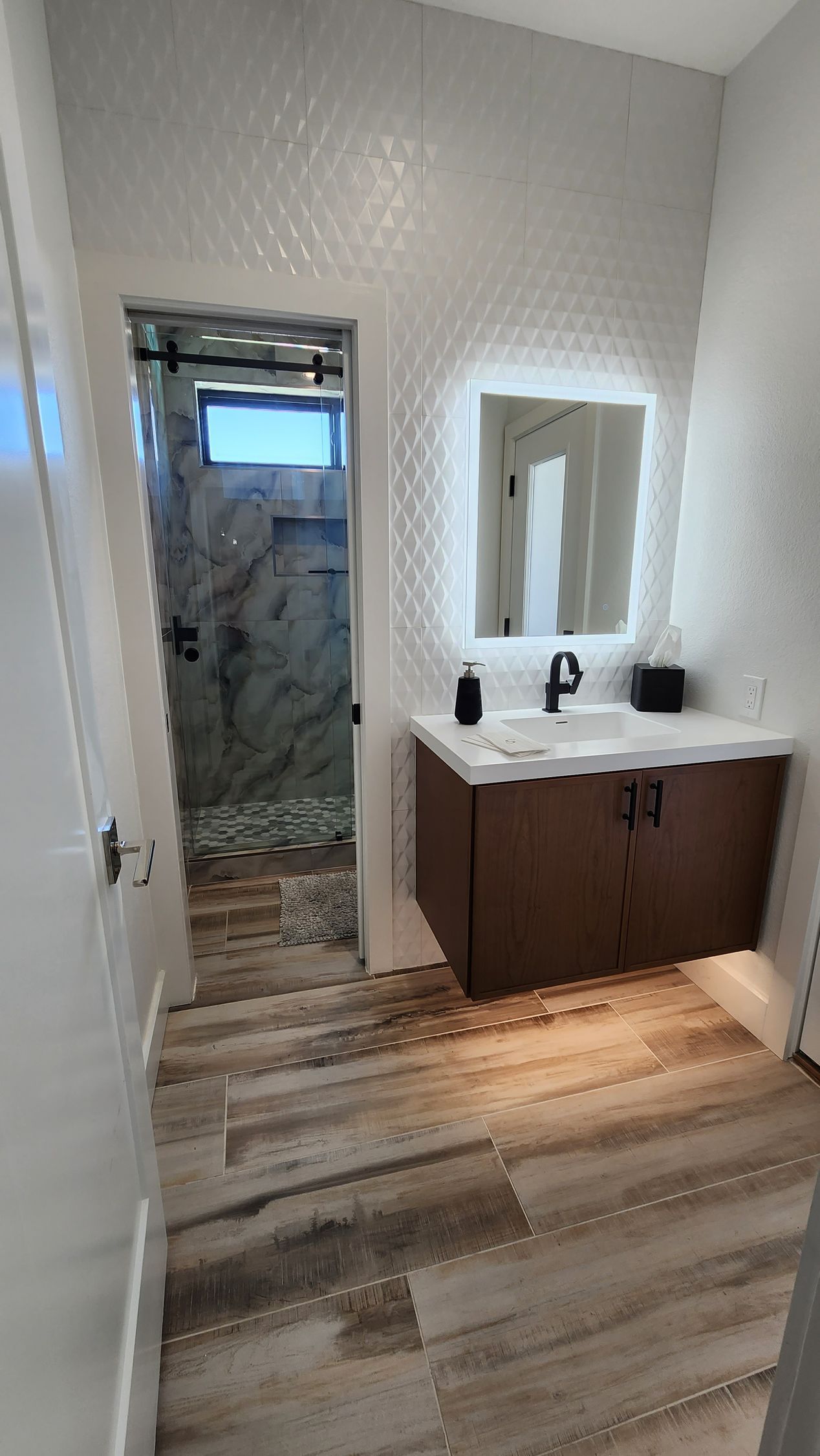Modern bathroom with white tiled walls, wood vanity, mirror, and shower visible through doorway