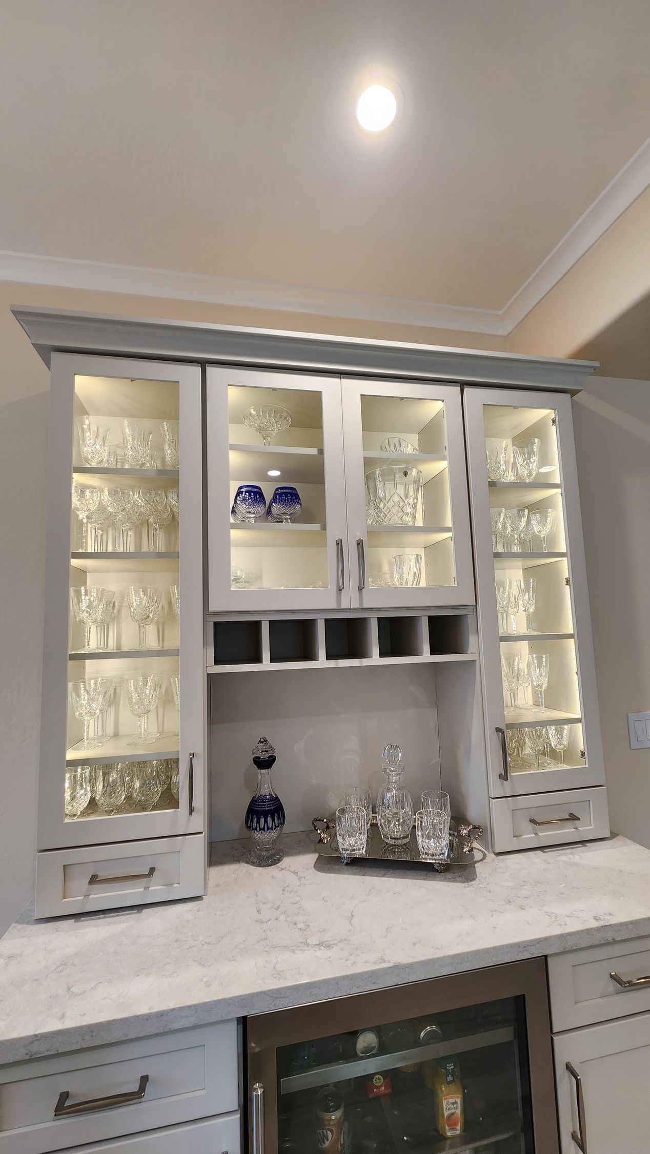 White kitchen hutch with glass doors and lit shelves displaying dishes, on a tiled countertop above an oven.