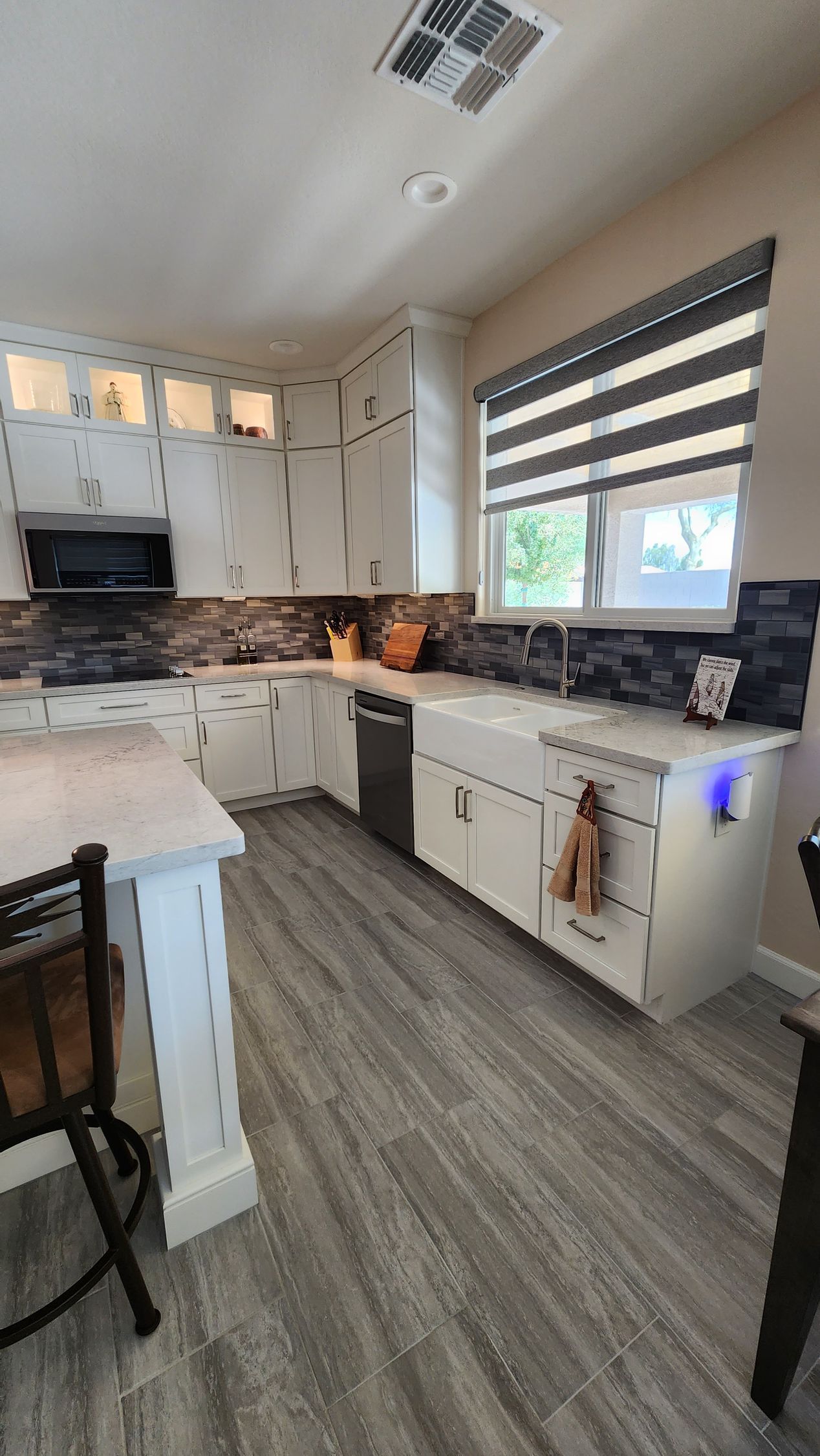 Bright modern kitchen with white cabinets, wood flooring, tiled backsplash, and a window with striped blinds.