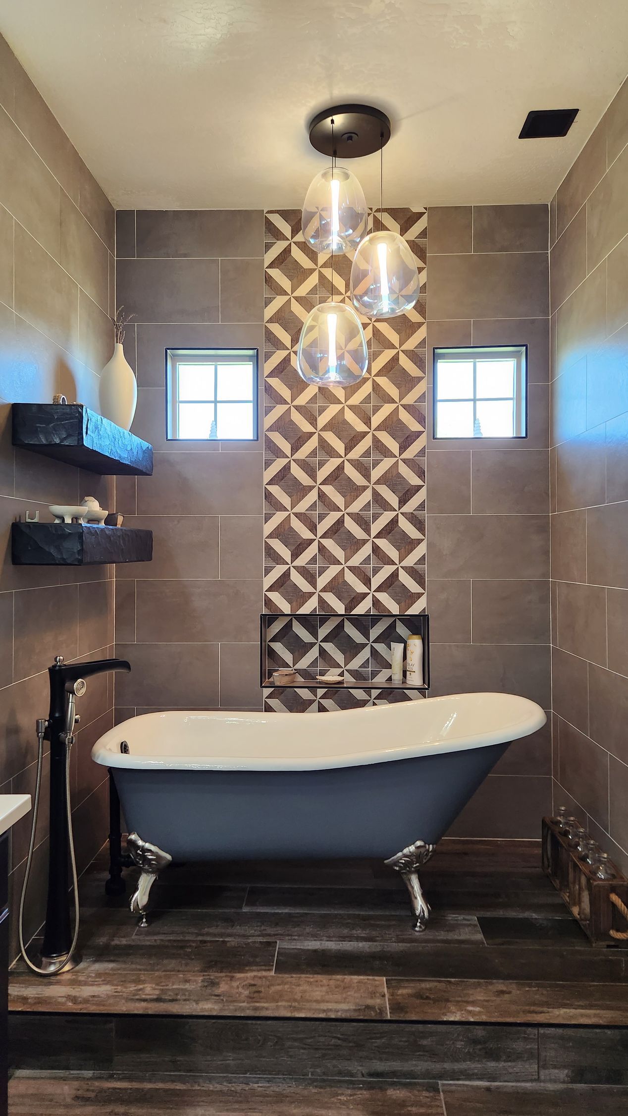 Modern bathroom with freestanding tub, patterned accent wall, pendant lights, and dark wood floor.