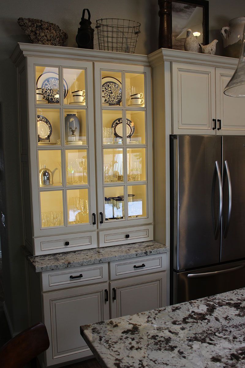 White kitchen cabinet with lit glass doors beside a stainless steel refrigerator and granite countertop