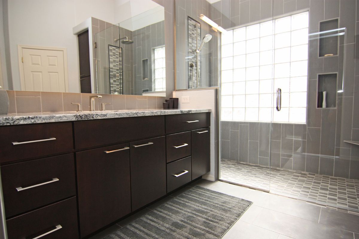 Modern bathroom with dark wood vanity, granite counter, large mirror, tiled shower, and gray floor mat