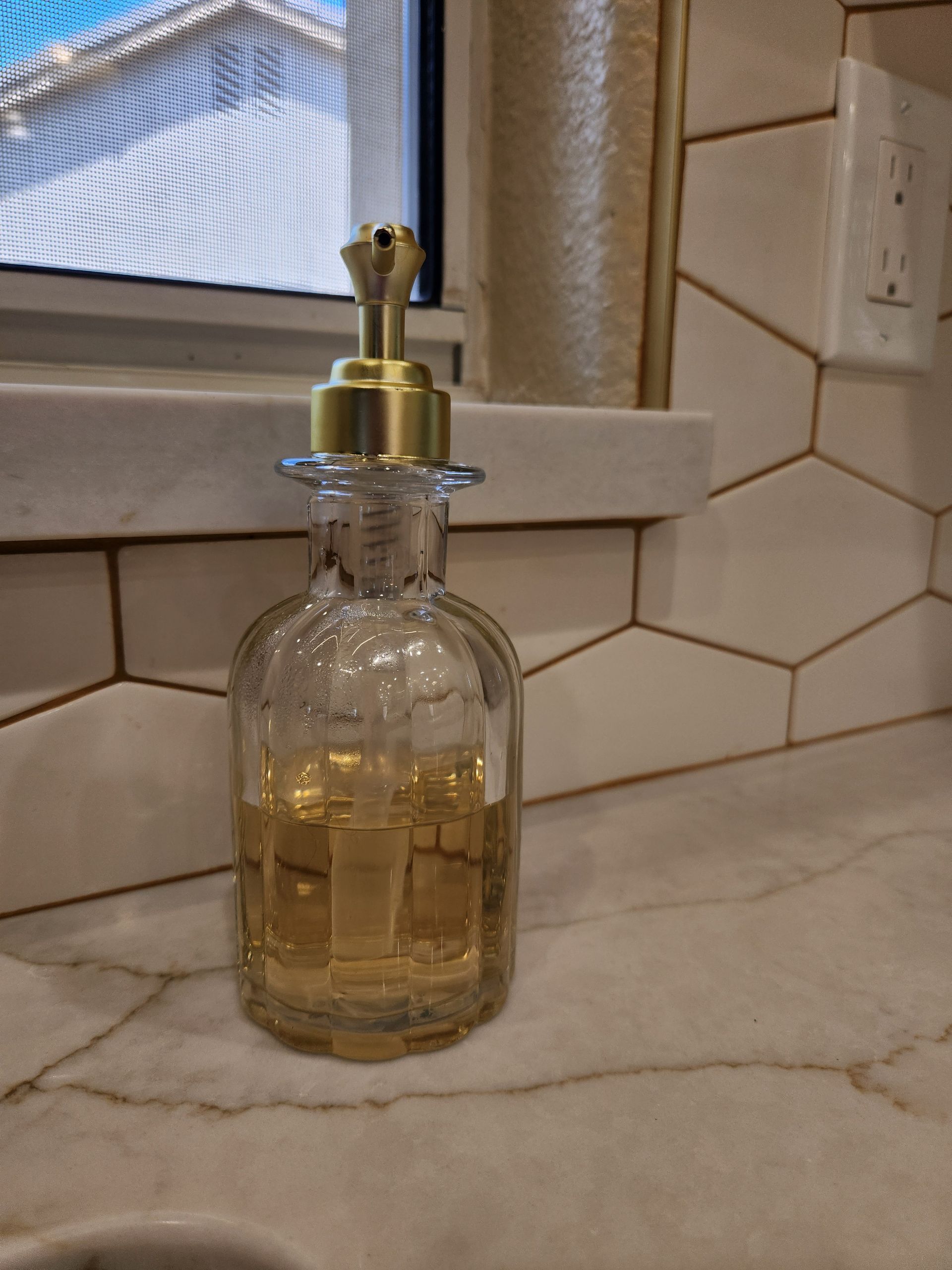 Clear glass soap dispenser on a bathroom counter by a window, filled with light amber liquid.