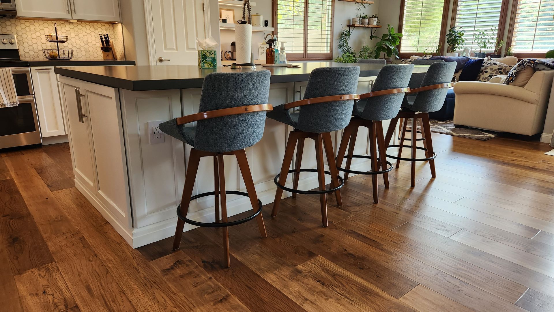 Modern kitchen with a large island, four blue stools, hardwood floors, and a living area in the background