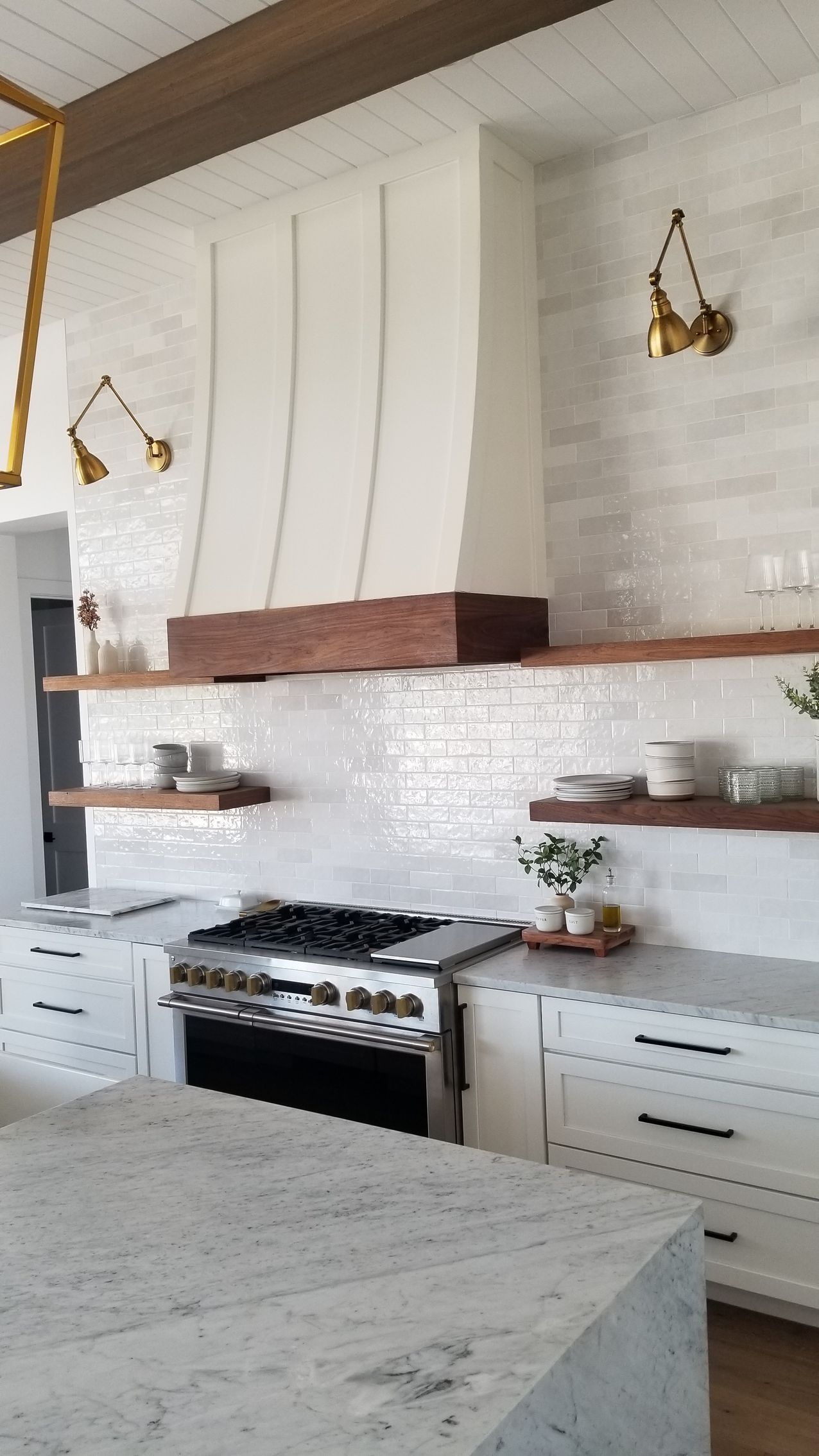 Bright white kitchen with marble island, stainless stove, wood shelves, and brass pendant lights.