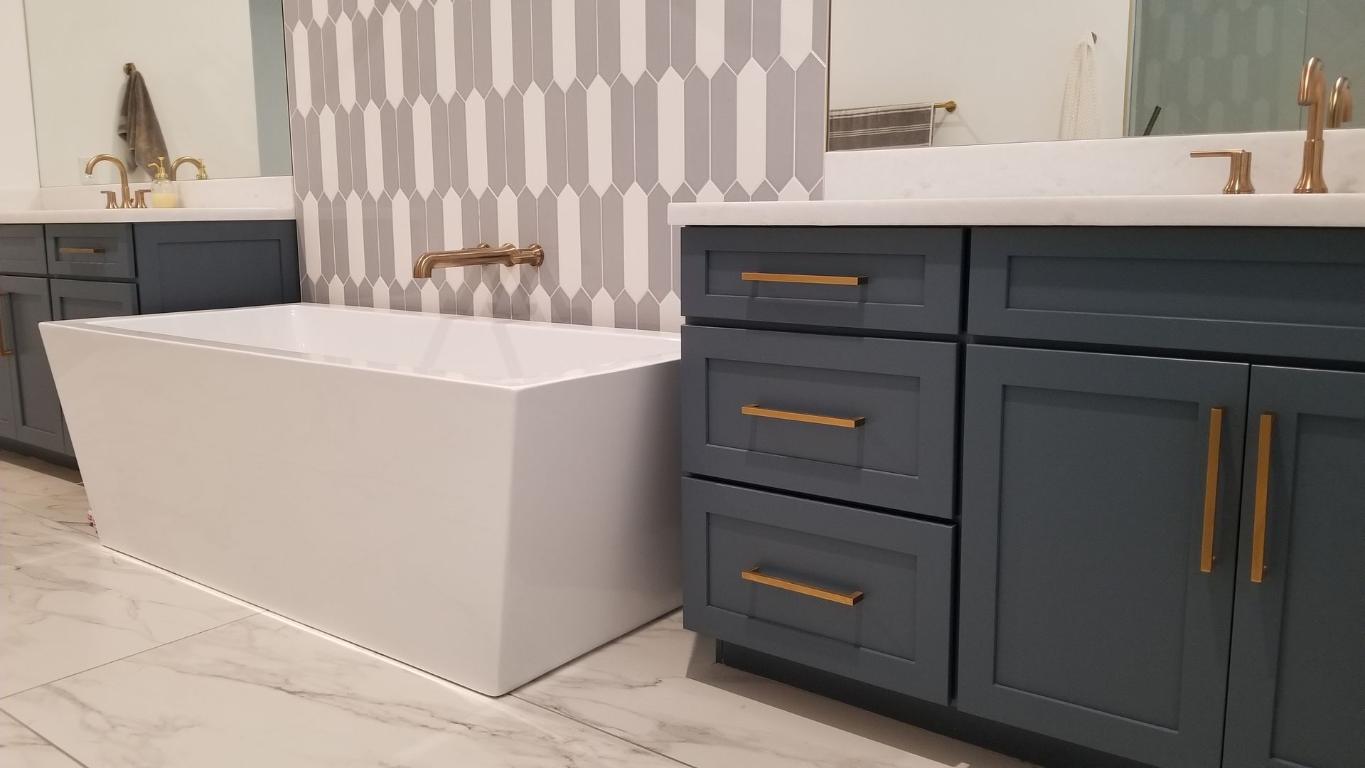 Modern bathroom with a white freestanding tub, navy vanity, brass fixtures, and patterned gray wall tile