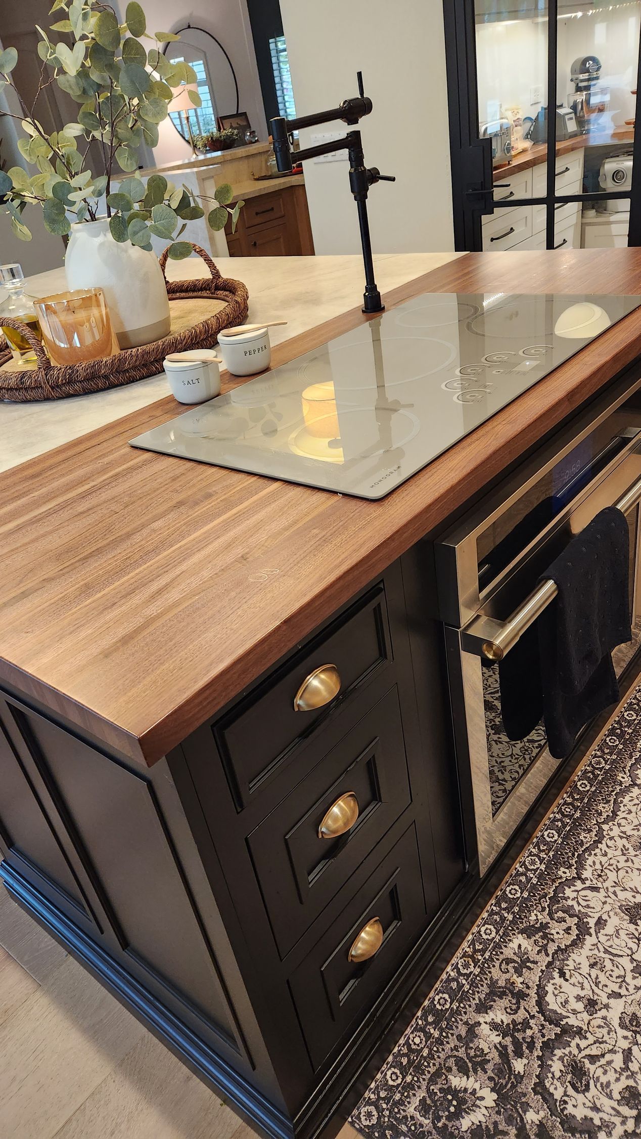 Kitchen island with glass cooktop, wood countertop, and black cabinets beside a patterned rug.