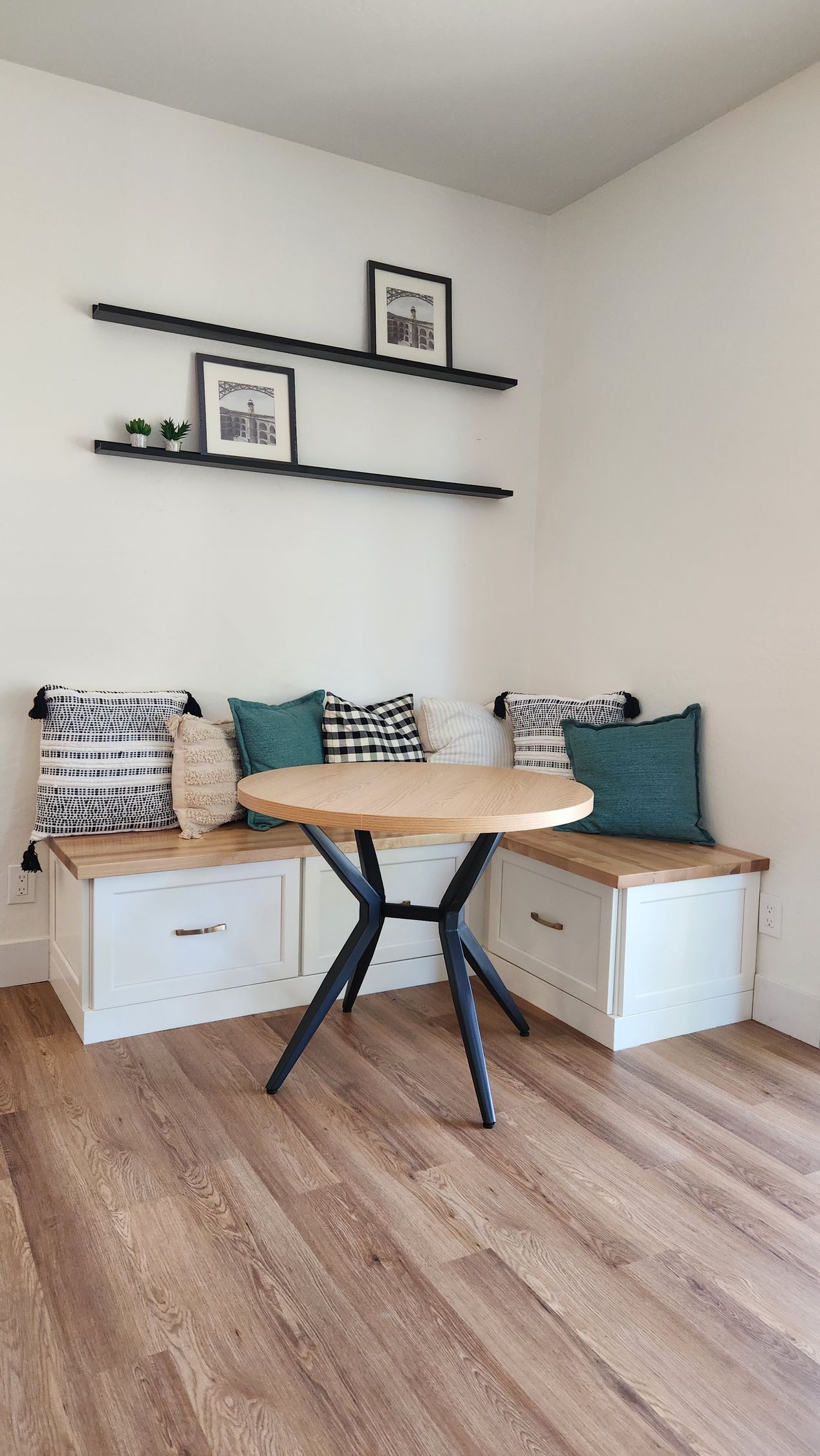 Small cozy corner with wooden bench seating, patterned cushions, floating shelves, and a small table on wood flooring