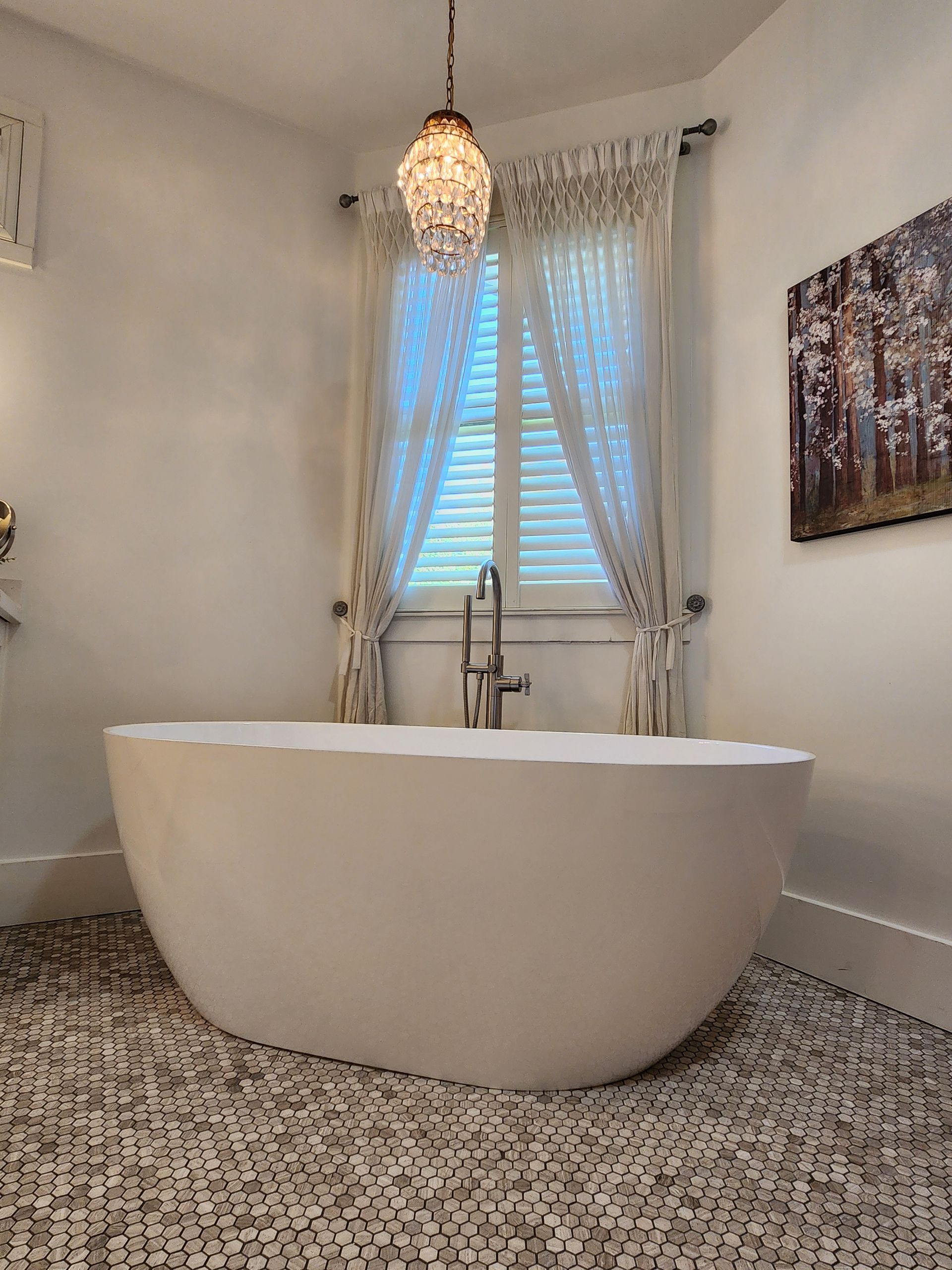 Modern bathroom with freestanding white tub, chandelier, and window with sheer curtains
