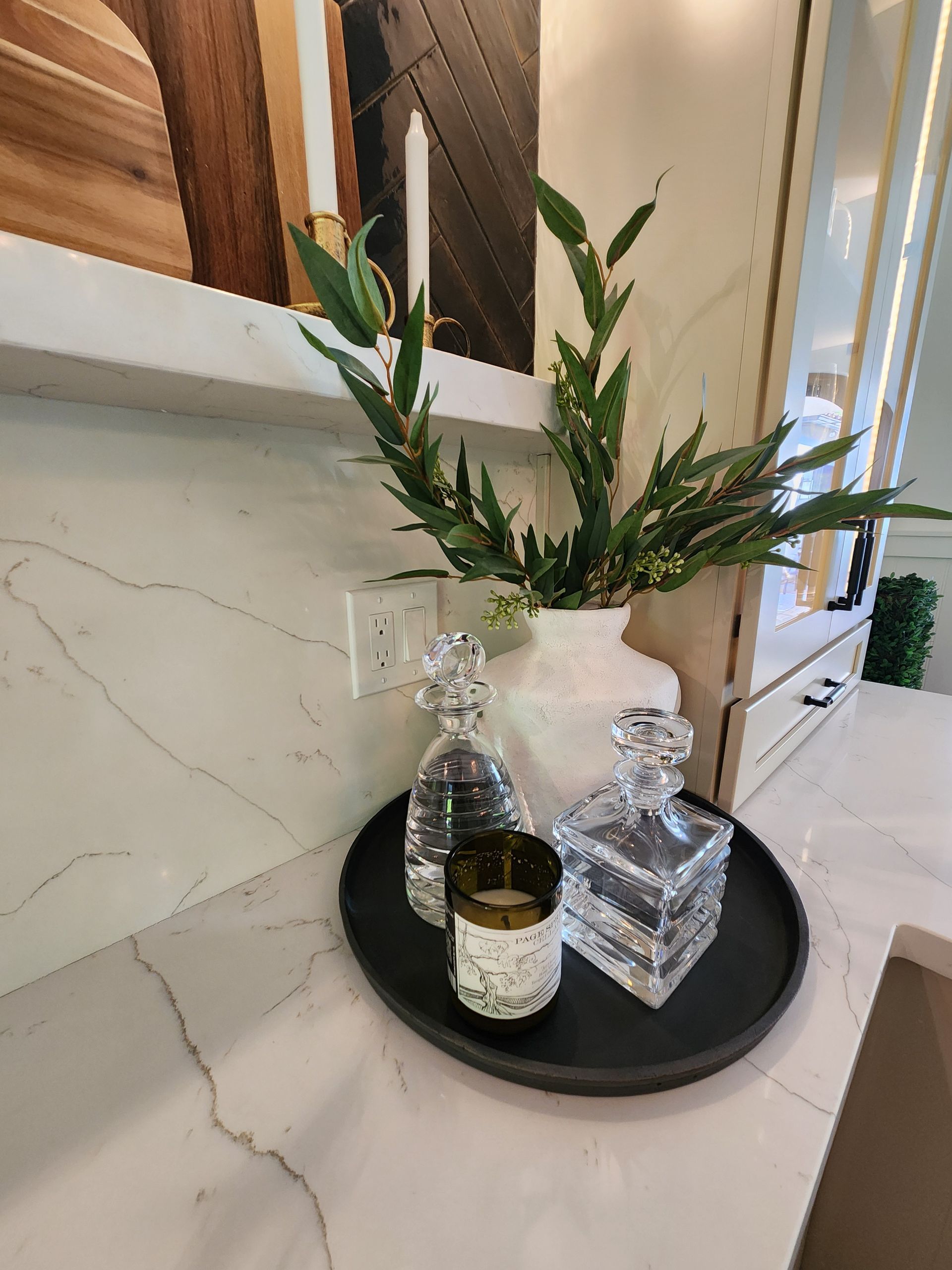 Decorative tray with glass bottles and greenery on a marble countertop against a mirror and wall.