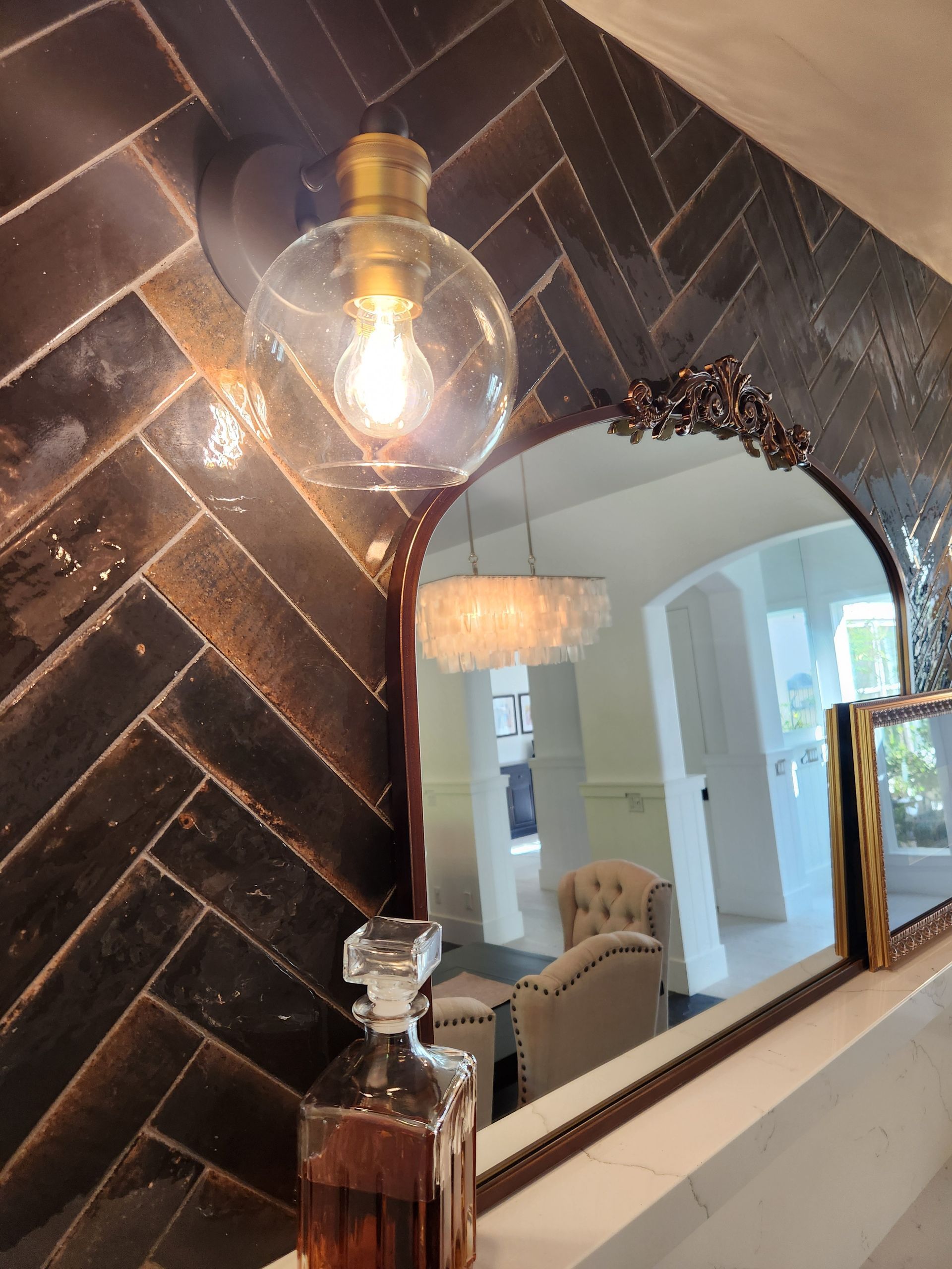 Dark tiled wall with glowing bulb above an ornate mirror reflecting a bright living room.