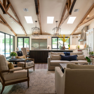 Bright living room with exposed beams, large windows, sofas, chairs, and a central fireplace.