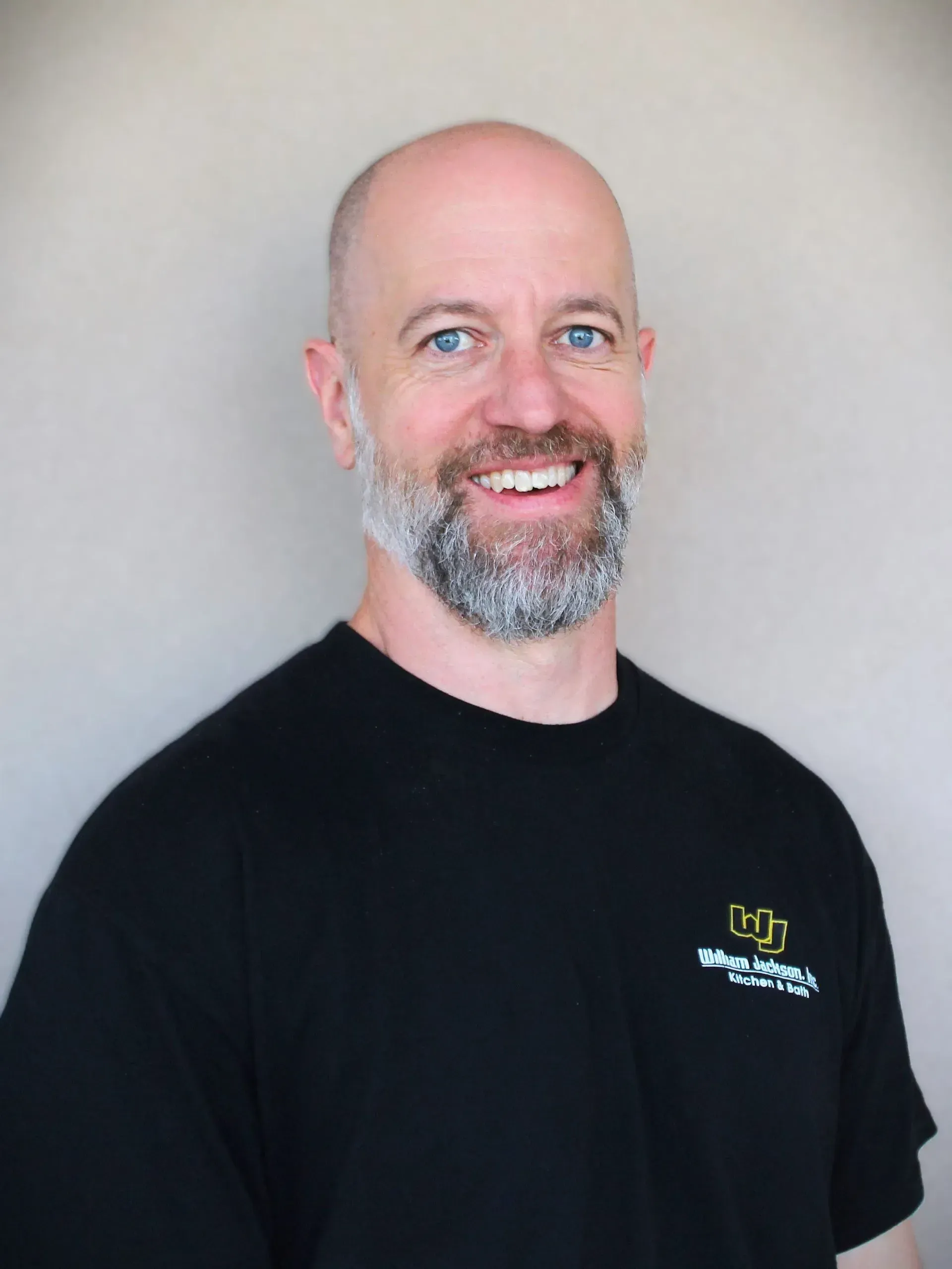 Smiling bald man with gray beard in a black T-shirt against a light gray background