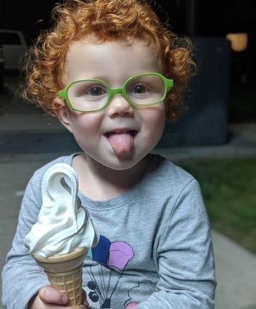 Child with curly red hair wearing green glasses, licking an ice cream cone outdoors at night.
