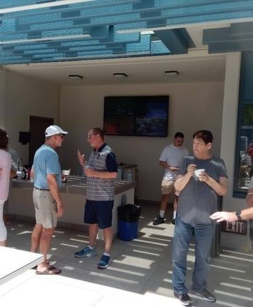 People chatting in a shaded outdoor bar area with a TV screen and stools.