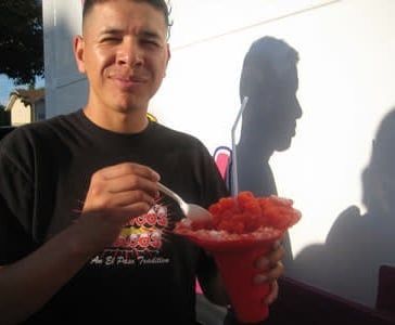 Man holding a cup of red shaved ice and spoon, standing in sunlight against a white wall