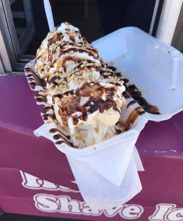 Shaved ice dessert in a foam container, topped with chocolate and caramel drizzle on a magenta table