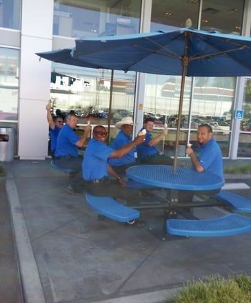 People in blue shirts sitting at a blue picnic table under an umbrella outside a building